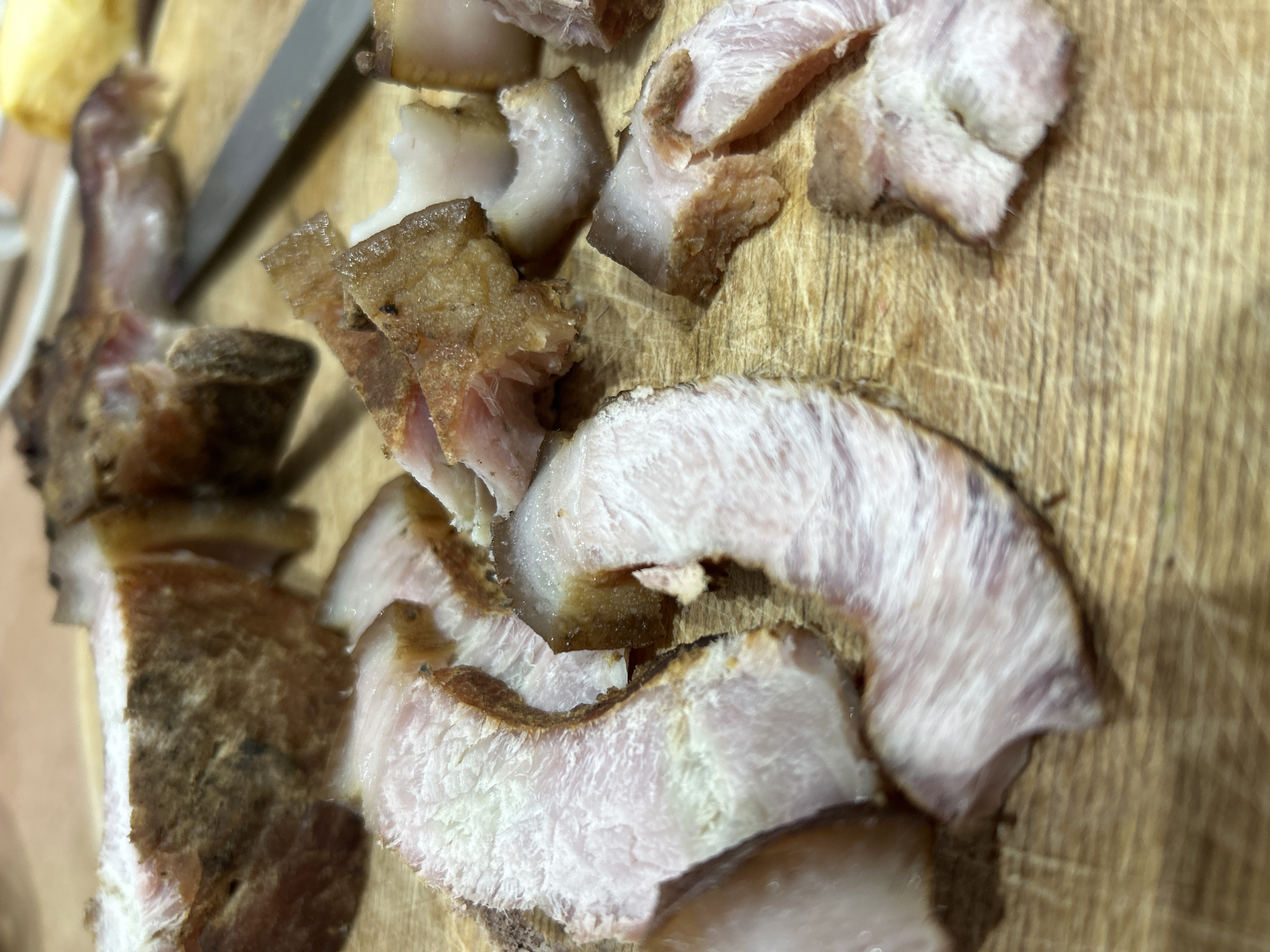 The image shows sliced pork belly on a wooden cutting board. A knife is visible in the background. The meat has a combination of white fat and pink muscle tissue, with a browned exterior. The pieces are irregularly shaped and scattered across the board. There is no evidence to determine if the location is in the city of Chaikovsky, Russia.