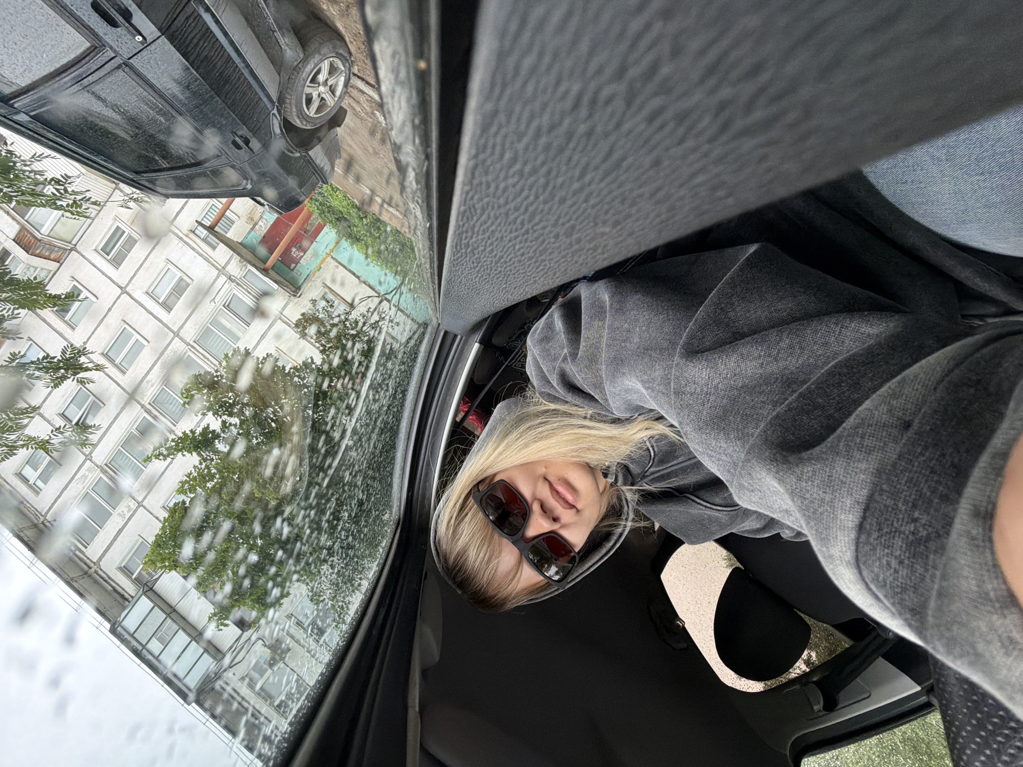 This vertical-format image captures a scene from inside a car on a rainy day in Chaikovsky, Russia. The image is originally rotated 90 degrees counter-clockwise.
The main subject is a young blonde woman, occupying the right side of the frame. She is looking upwards and slightly to the left, wearing dark, square-framed sunglasses and a grey hooded sweatshirt or jacket. Her expression is neutral, with her lips slightly parted. The interior of the car is visible around her, including dark fabric materials of the seats and headliner.
Outside the car window, on the left side of the image, raindrops are clearly visible on the glass, indicating current or recent rain. A dark-colored car, possibly an SUV or jeep, is parked nearby, its wet surface reflecting the overcast sky. In the background, several multi-story residential buildings with multiple windows are seen, characterized by light-colored facades. Green trees and foliage are interspersed among the buildings and the parked car, also appearing damp from the rain. The overall setting suggests a residential area within an urban or suburban environment. The weather appears to be overcast, contributing to a muted lighting. There is no discernible text visible in the image.