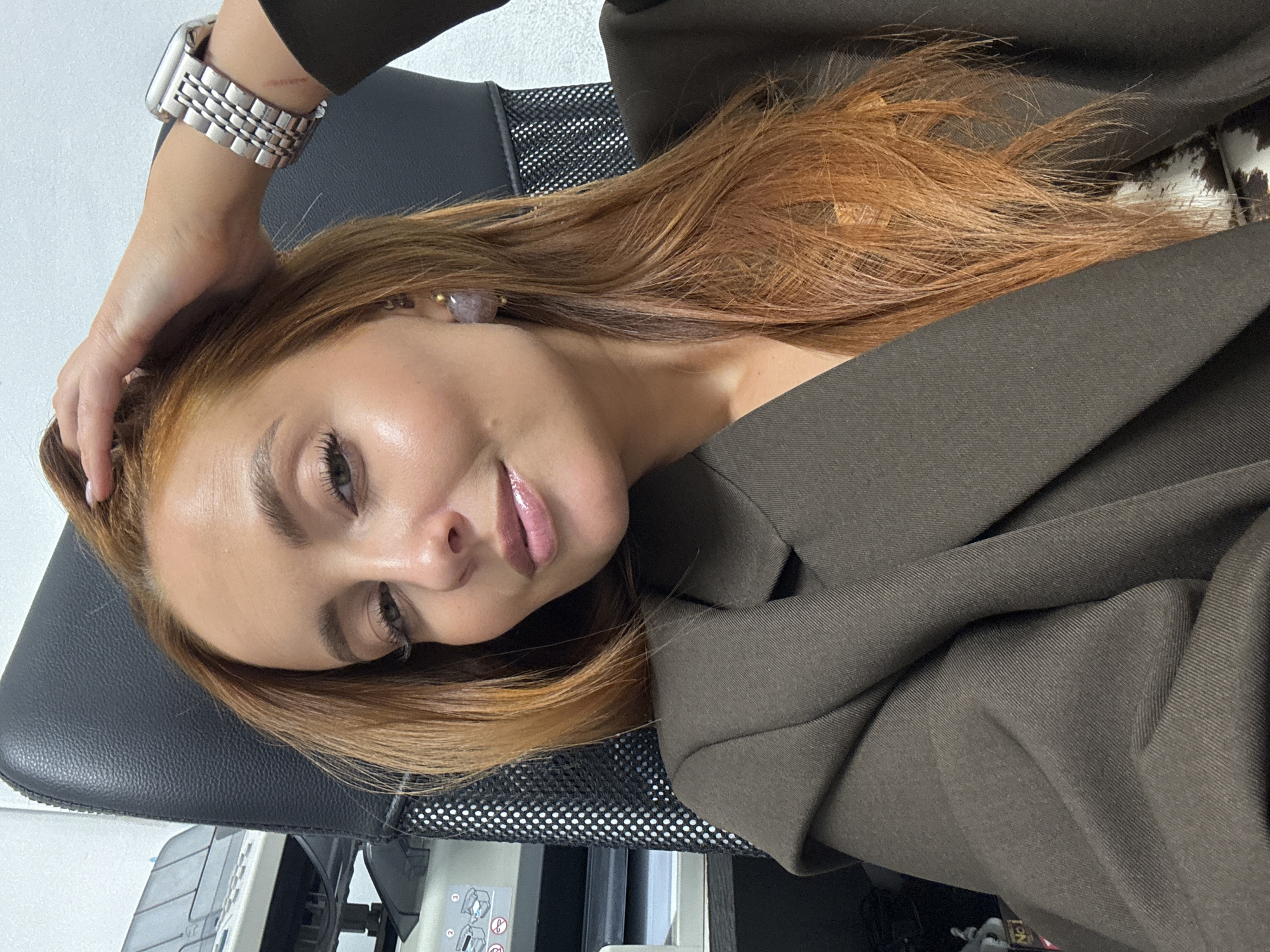 A close-up portrait features a woman with long, reddish-brown hair, light skin, and natural-looking makeup, including glossy pink lips. She is lying or reclining against a black office chair, looking towards the camera with a relaxed, soft smile and partially closed eyes. Her posture suggests a calm and serene mood.
She is dressed in a dark, possibly olive-green, blazer or jacket over a dark top. On her left wrist, she wears a silver-colored smartwatch with a metal link band. Her right hand is gently placed on her head, lightly touching her hair. A light purple or pinkish gemstone earring is visible in her right ear.
The setting appears to be an indoor office environment. The background includes parts of the black mesh backrest of the chair she is leaning on, and out-of-focus office equipment such as a printer or scanner. A label with "No. 1" is partially visible on this equipment. Below it, a white sticker shows two simplified stick figures, one standing with an 'X' mark and one sitting with a checkmark, likely indicating "do not stand, sit" instructions. Further back, a pattern resembling white and brown animal print (possibly cowhide) is faintly discernible. The lighting is soft and even, highlighting her features. The image itself is tilted, giving a dynamic angle.