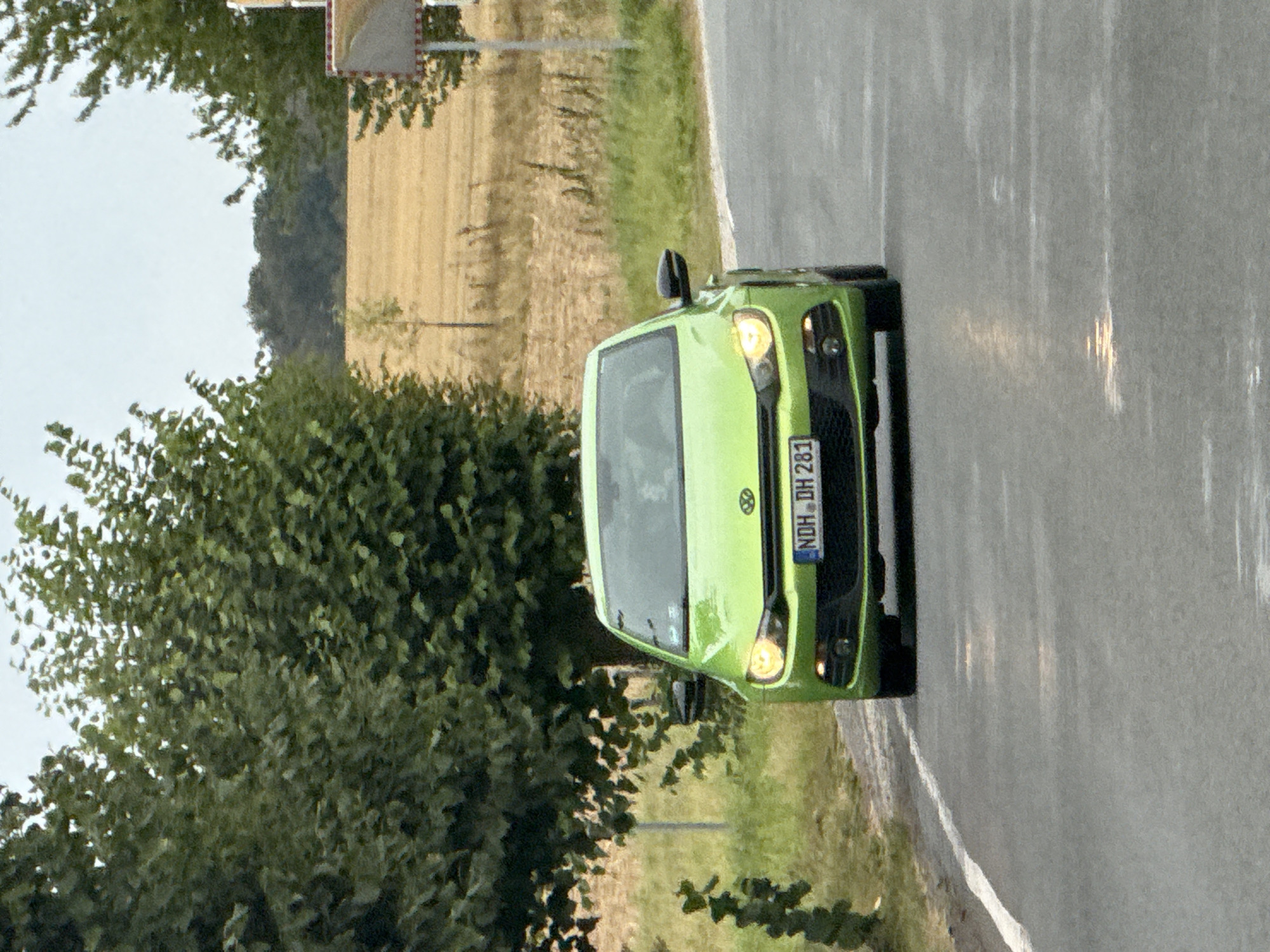 A bright green Volkswagen Scirocco is driving on a wet asphalt road. The car's headlights are on, illuminating the road ahead. The car has a license plate that reads "NDH DH 281". The car is surrounded by lush green trees and vegetation. In the background, there is a field of dry grass, suggesting a rural setting. A speed limit sign is visible on the right side of the road, indicating the location as Hohenstein, Germany. The sky is overcast, hinting at potential rain. The image captures a moment of transit, with the car the central subject against a backdrop of nature.
