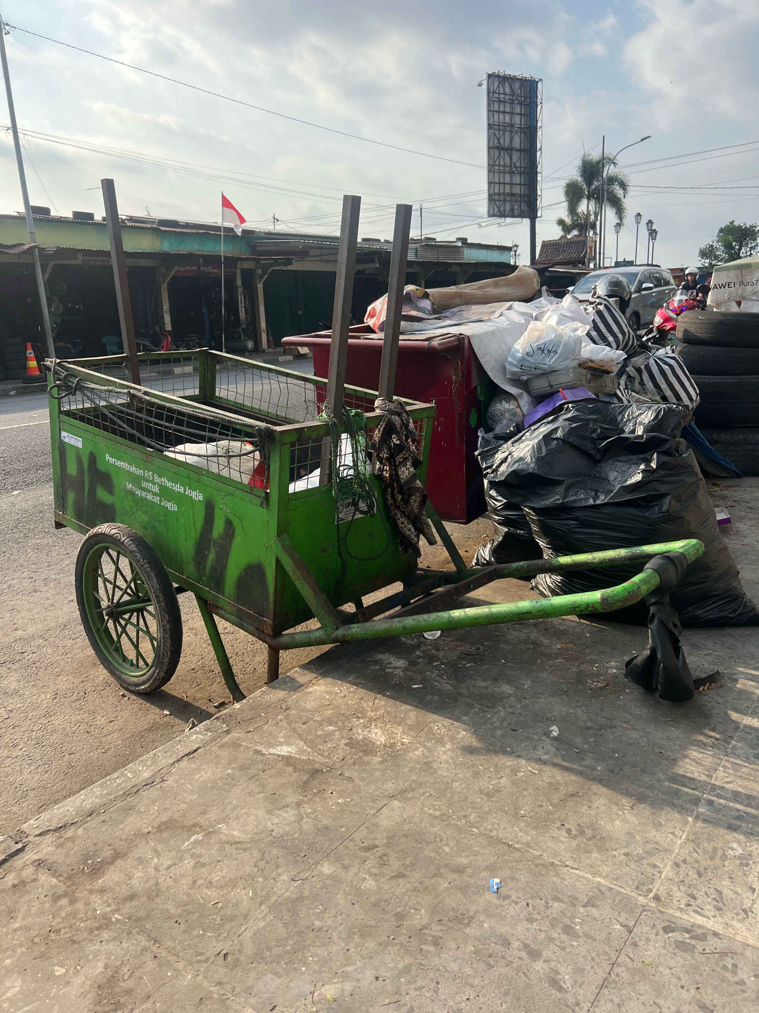A green metal cart with a single wheel sits on a concrete sidewalk next to a pile of trash bags and other debris. The cart has "Persembahan RS Bethesda Jogja untuk Masyarakat Jogja" written on its side, which translates to "Gift from Bethesda Hospital Yogyakarta for the people of Yogyakarta." In the background, there are market stalls with a red and white Indonesian flag flying and a large billboard. A motorcycle and several people are visible in the distance. The scene appears to be an outdoor area in Yogyakarta, Indonesia, likely a street or market.