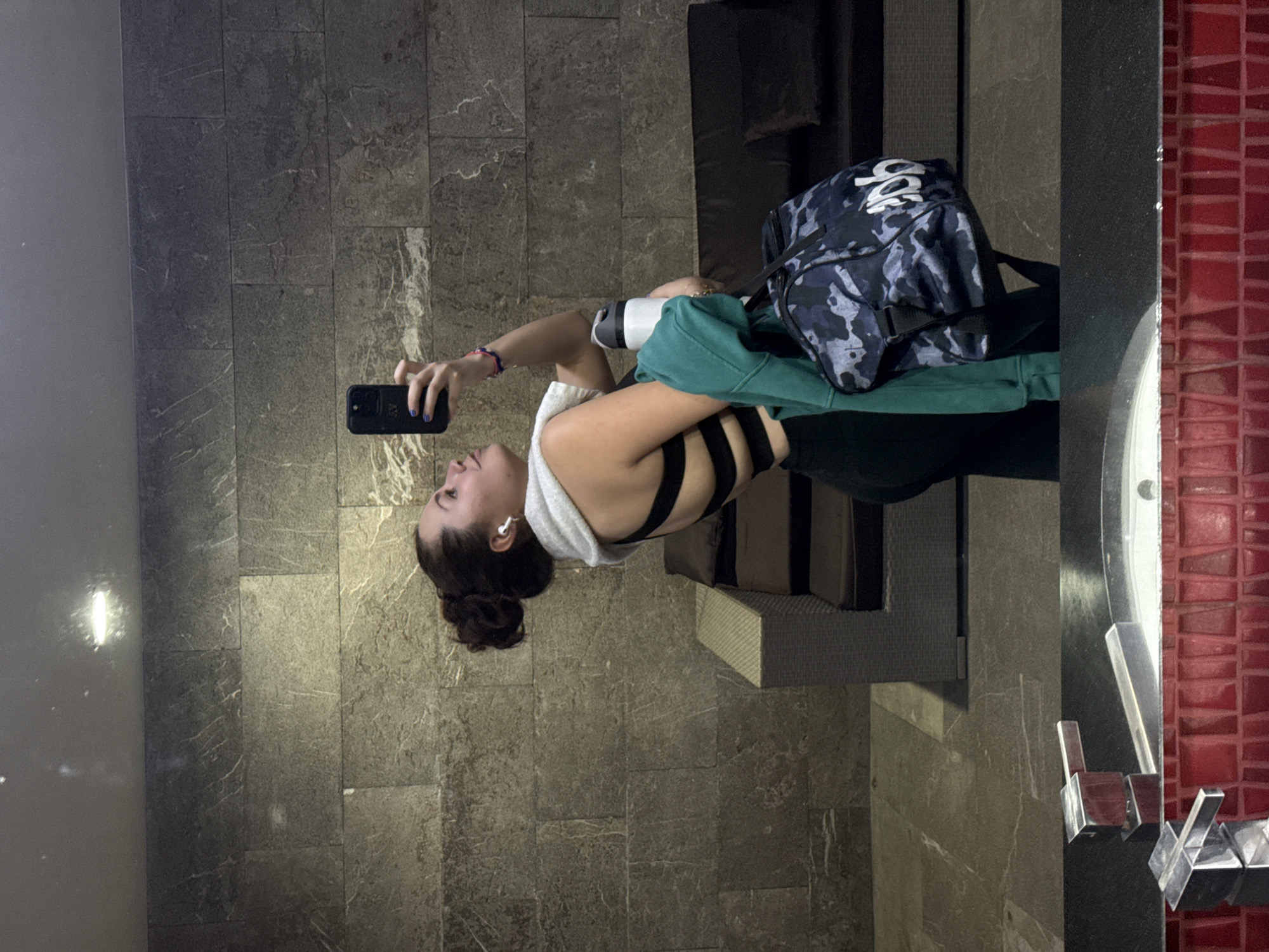 A young woman is standing in front of a mirror, holding a black smartphone in her left hand and looking at the screen. She is wearing a black sports bra with multiple straps across her back and a white hooded sweatshirt. Her hair is pulled back in a bun. She also has what appears to be wireless earbuds in her ears. Her fingernails on her left hand appear to be painted a dark blue.
Behind her is a bench with a blue and gray camouflage-patterned duffel bag and a green hooded sweatshirt on it. The floor and walls are covered in gray tiles. To the right of the frame, there is a red tiled wall and part of a sink with metal fixtures.
Based on the architectural details and the overall aesthetic, it is likely that the location is a modern gym or spa in Mexico City, Mexico.
