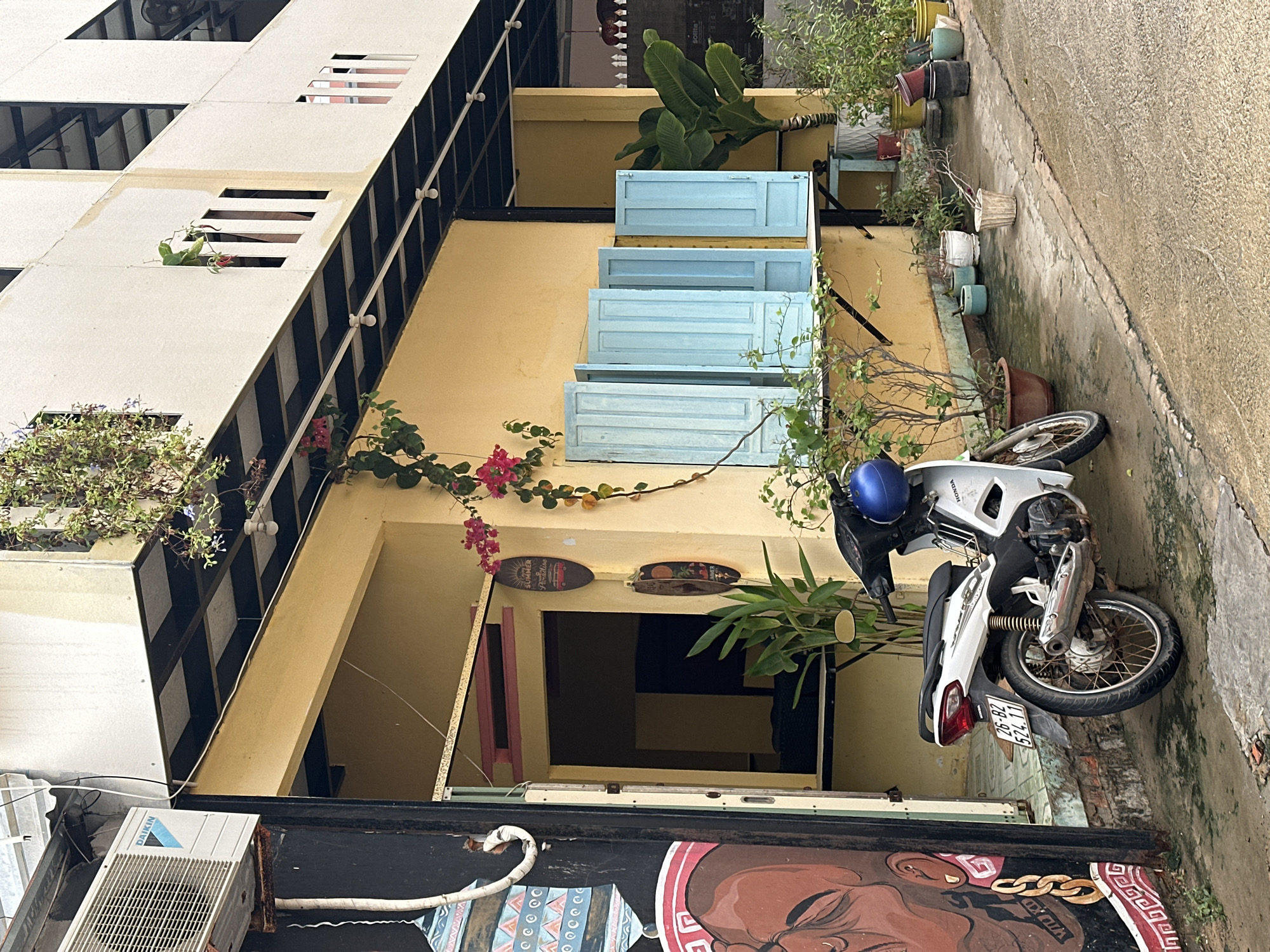 A street scene in Phu Thuy Ward, Vietnam, shows a building with yellow walls and a white and black facade. Three blue shutters adorn the building. A white Honda motorcycle with a blue helmet sits parked on the sidewalk. Planters line the edge of the sidewalk. An air conditioning unit is attached to the building. A mural of a face is visible on the bottom of the frame.