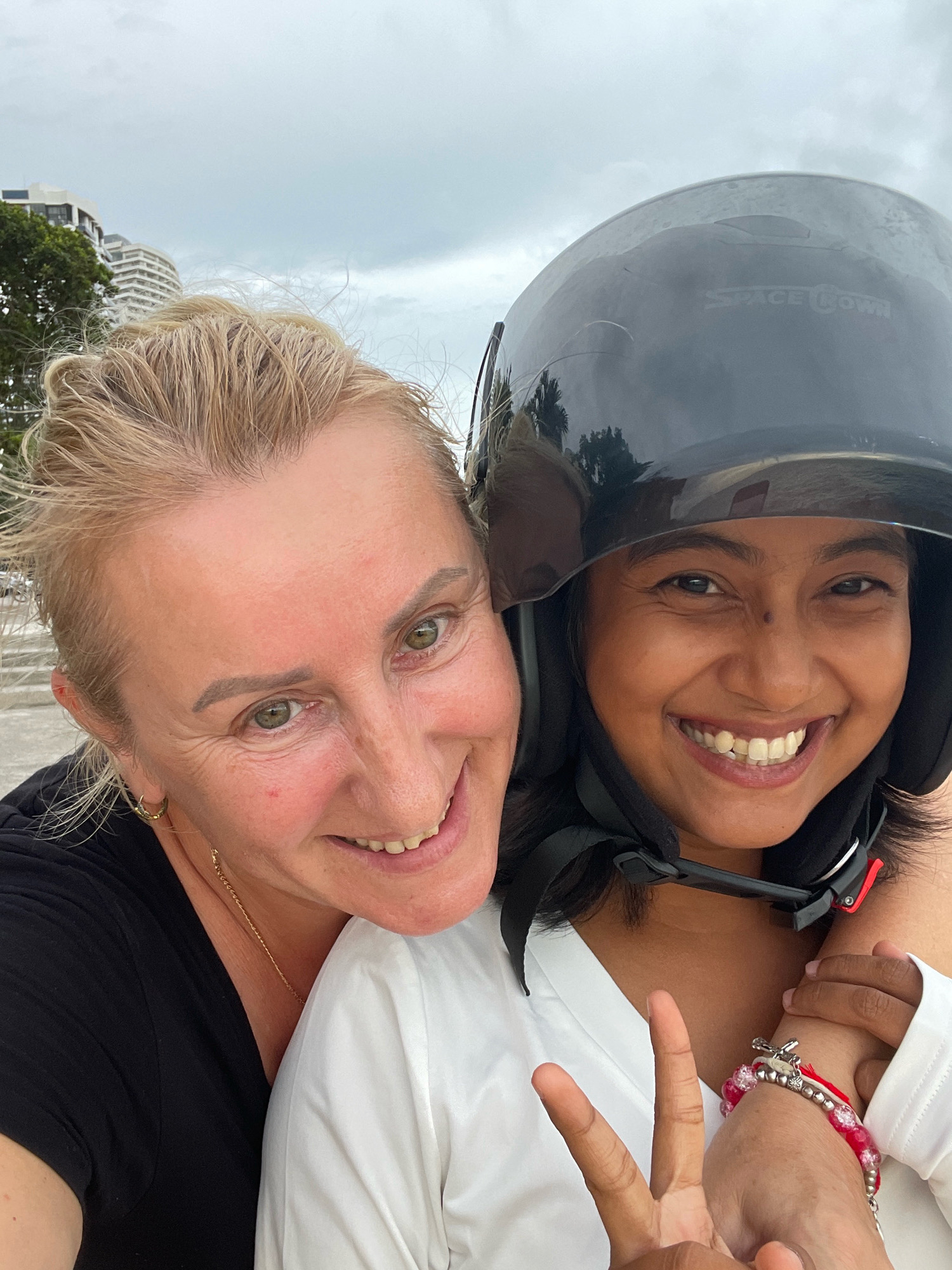 In a selfie, two women smile with their heads close together. The woman on the left, with blonde hair and fair skin, wears a black shirt and gold hoop earrings. She is looking towards the camera with her eyes open. The woman on the right, with dark hair and tan skin, wears a white shirt and a black helmet. Her eyes are also open and she is smiling broadly. She is making a peace sign with her right hand, which is adorned with a red beaded bracelet. The background shows a cloudy sky and buildings, suggestive of a city environment. The presence of buildings and a cloudy sky may indicate an urban setting like Pattaya City, Thailand.