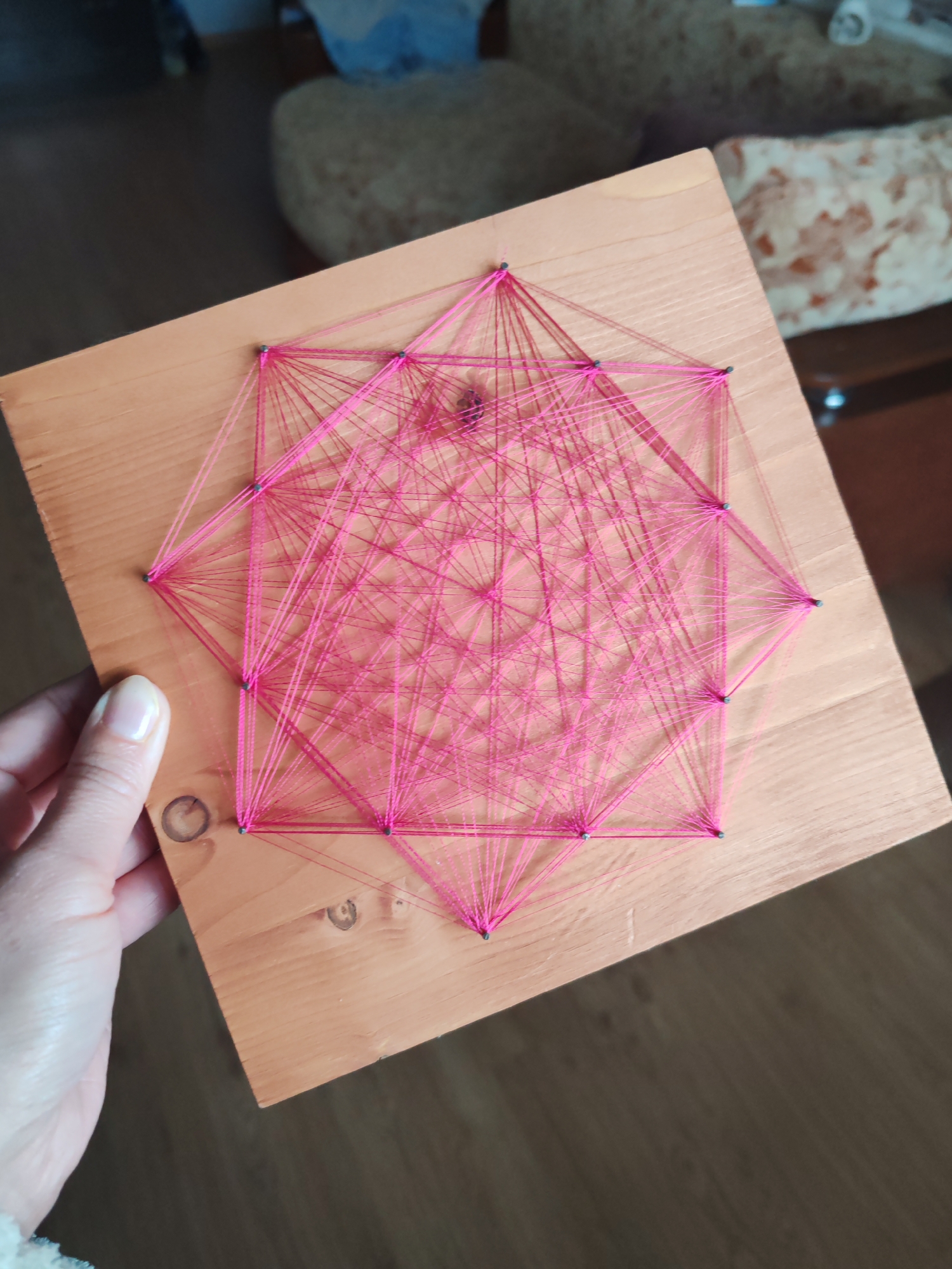 A close-up shot depicts a square piece of light-colored wood on which a geometric design has been crafted using pink thread. Small, dark pins are visible at the corners of the design, holding the thread in place. A human hand with fair skin and neatly manicured nails holds the wooden piece. The background is slightly blurred, featuring a glimpse of furniture including a floral-patterned cushion or sofa. The location and city are not ascertainable from the provided image alone.