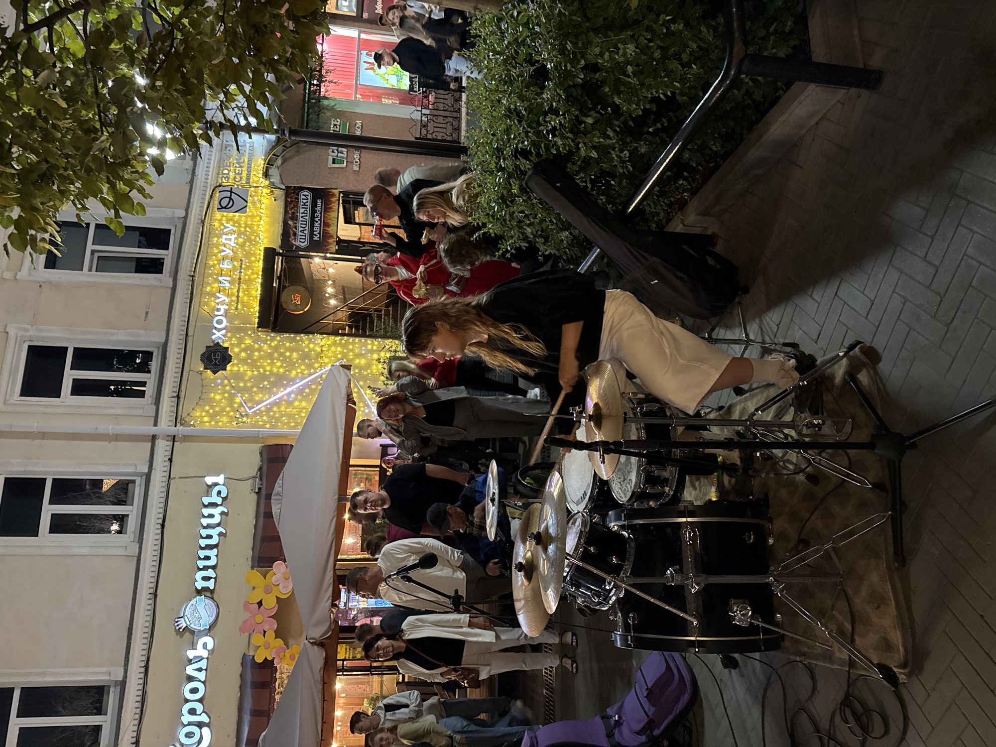 The image captures an evening street scene in Kazan, Russia, featuring a live music performance. In the foreground, a young woman with long blonde hair is seated and playing a drum set. She is wearing a black long-sleeved shirt and white pants. The drum set, consisting of multiple drums and cymbals, is positioned on a rug on the cobblestone street. Several microphones are visible, including one positioned near a seated man in a blue shirt and a black cap who appears to be part of the band.
Behind the performers, a crowd of people is gathered, some watching the performance, others socializing. They are standing on the sidewalk in front of various establishments. Visible signage in Russian indicates a pizza place called "Король Пиццы" (King of Pizza) and another establishment with the text "Хочу и Буду" (I Want and Will Be). The buildings are illuminated with strings of yellow lights, creating a festive atmosphere. Some buildings have awnings or umbrellas set up outside. The overall environment suggests a lively urban setting with ongoing entertainment.