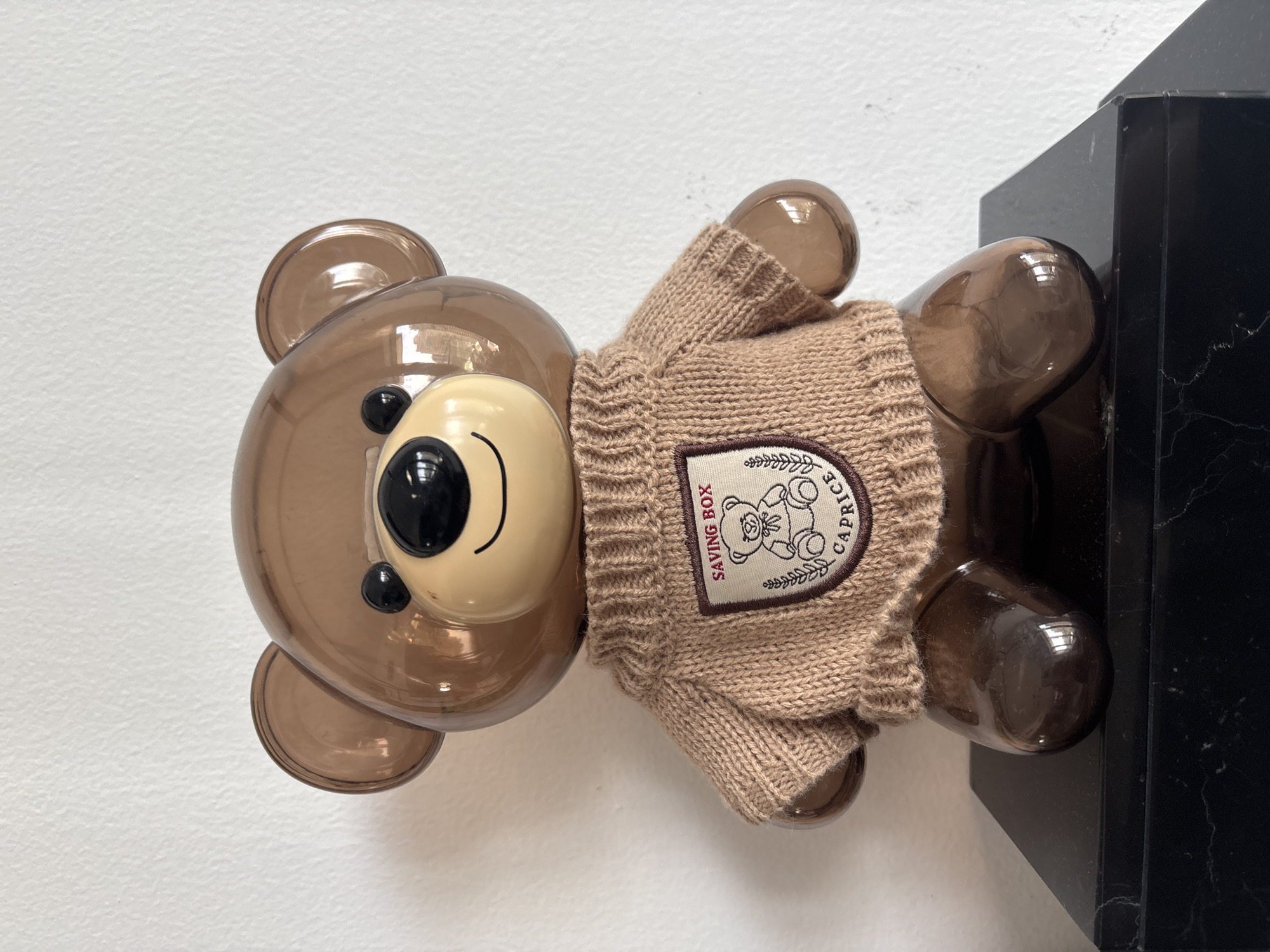 The image shows a close-up, eye-level, frontal view of a translucent brown teddy bear-shaped savings box sitting on a black marble surface. The teddy bear is wearing a knitted brown sweater with a patch on the chest. The patch has a drawing of a teddy bear and the words "SAVING BOX" and "CAPRICE". The background is a plain white wall. The scene is indoors and appears to be well-lit. There is no indication of the location being Dubai, United Arab Emirates.