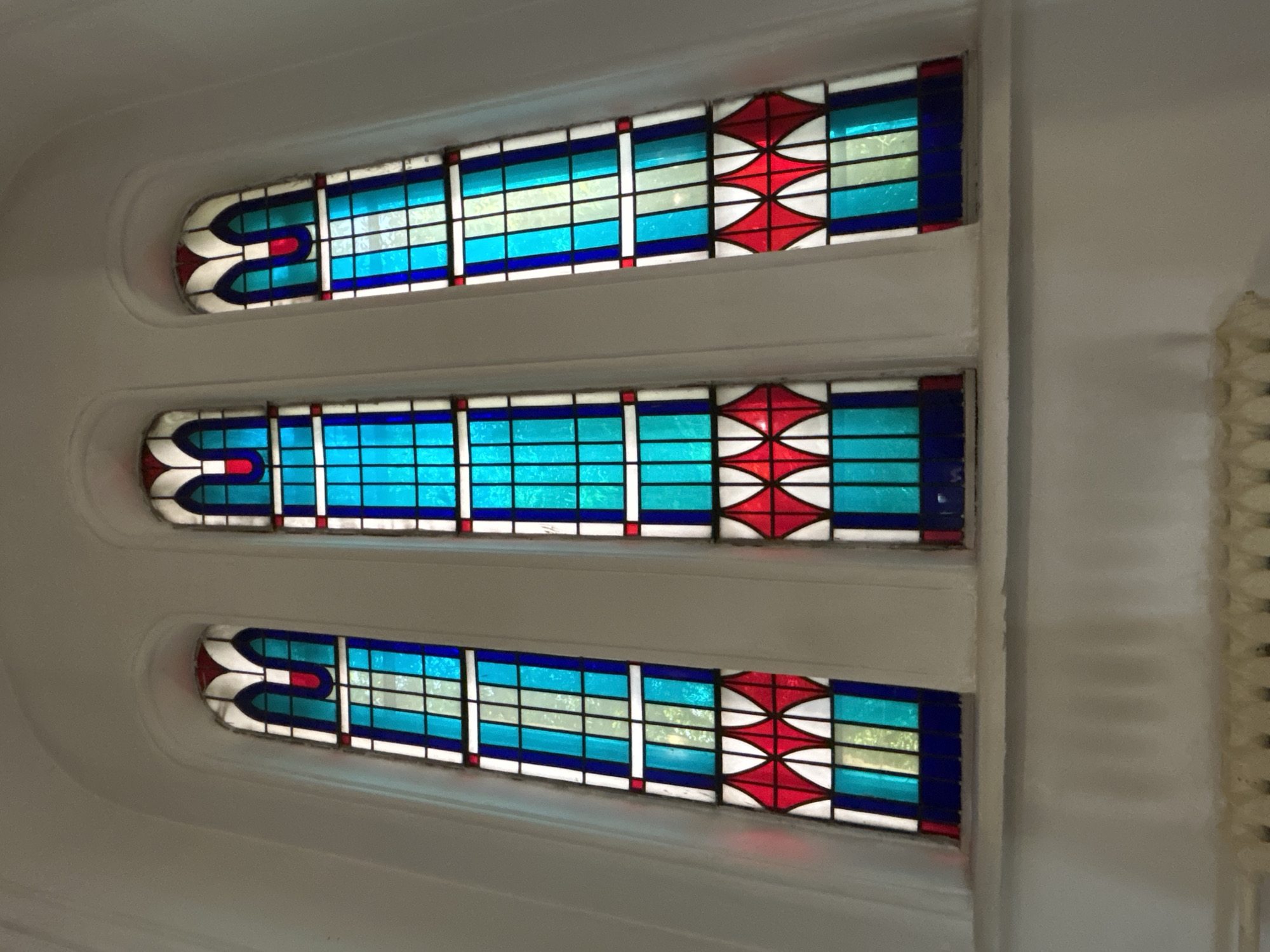 The image shows three tall, narrow stained-glass windows with arched tops, set into a white, curved wall. The windows feature a repeating geometric pattern of blue, green, white, and red glass. The bottom of each window has a diamond pattern in red and white, with blue and green rectangles above. The wall appears to be part of an interior space, with a radiator visible at the bottom of the frame. The lighting suggests daytime, with light filtering through the stained glass. The overall impression is of a religious or historical building. This image is a photograph of the stained-glass windows of the Tomsk Regional Museum of Local Lore.