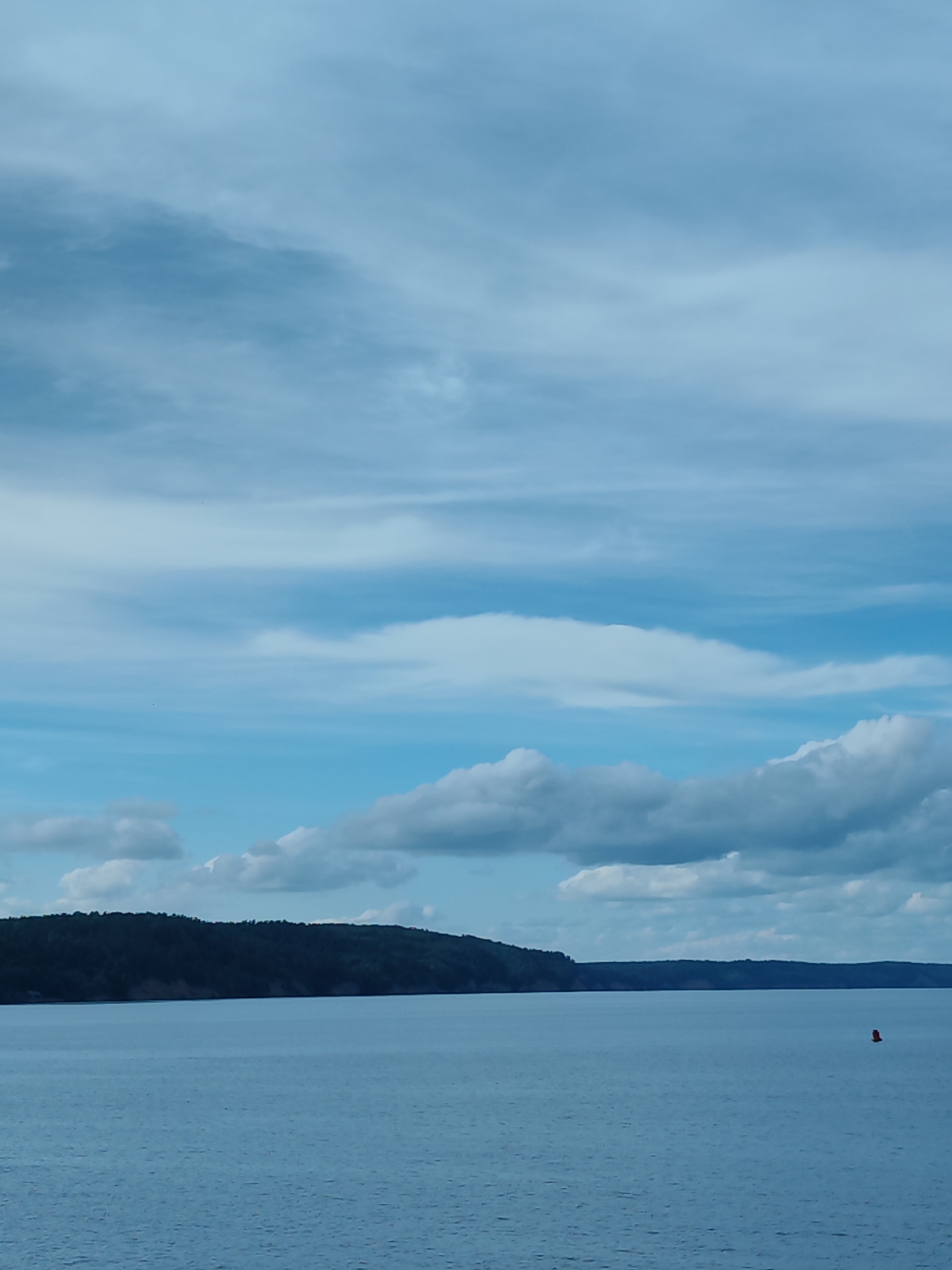 The image depicts a wide expanse of calm, blue water under a sky filled with layered clouds of varying shades of blue and white. A dark, tree-covered shoreline stretches across the horizon, creating a stark contrast with the water and sky. In the foreground, the water is a deep blue, with subtle ripples indicating gentle movement. Towards the right side of the frame, a small, red buoy is visible in the water. The overall scene conveys a sense of tranquility and vastness.