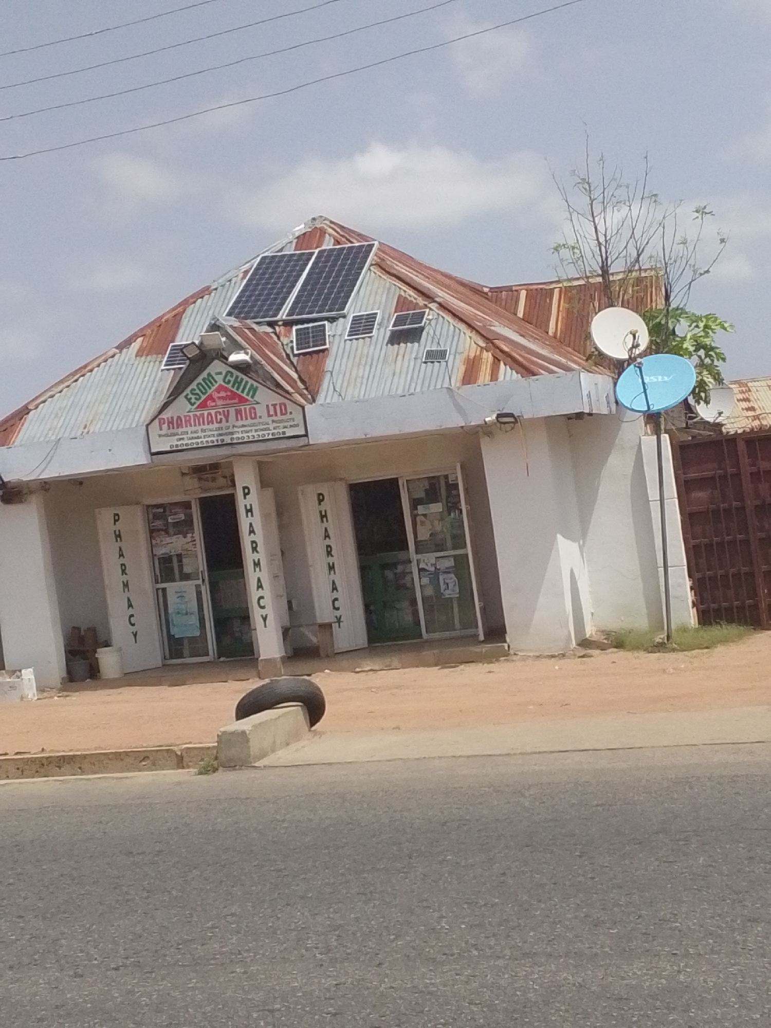 The image depicts the exterior of a pharmacy named "ESOM-CHIN PHARMACY NIG. LTD." in Jalingo, Nigeria. The pharmacy is a single-story building with a corrugated metal roof that shows signs of rust and age. Several solar panels are installed on the roof, indicating an attempt to utilize renewable energy. Two satellite dishes are also visible, one white and one blue, mounted on poles near the building. The building's facade is painted white, and there are large glass windows and doors, allowing a partial view of the interior which appears to be stocked with various items. The signage above the entrance clearly displays the pharmacy's name and a phone number. The building is situated on a dirt road, with a paved road in the foreground. A discarded tire rests on a concrete block near the roadside. The environment appears to be a commercial area within Jalingo, characterized by the presence of businesses and street infrastructure. The sky is cloudy, suggesting a daytime scene. There are no people visible in the image.