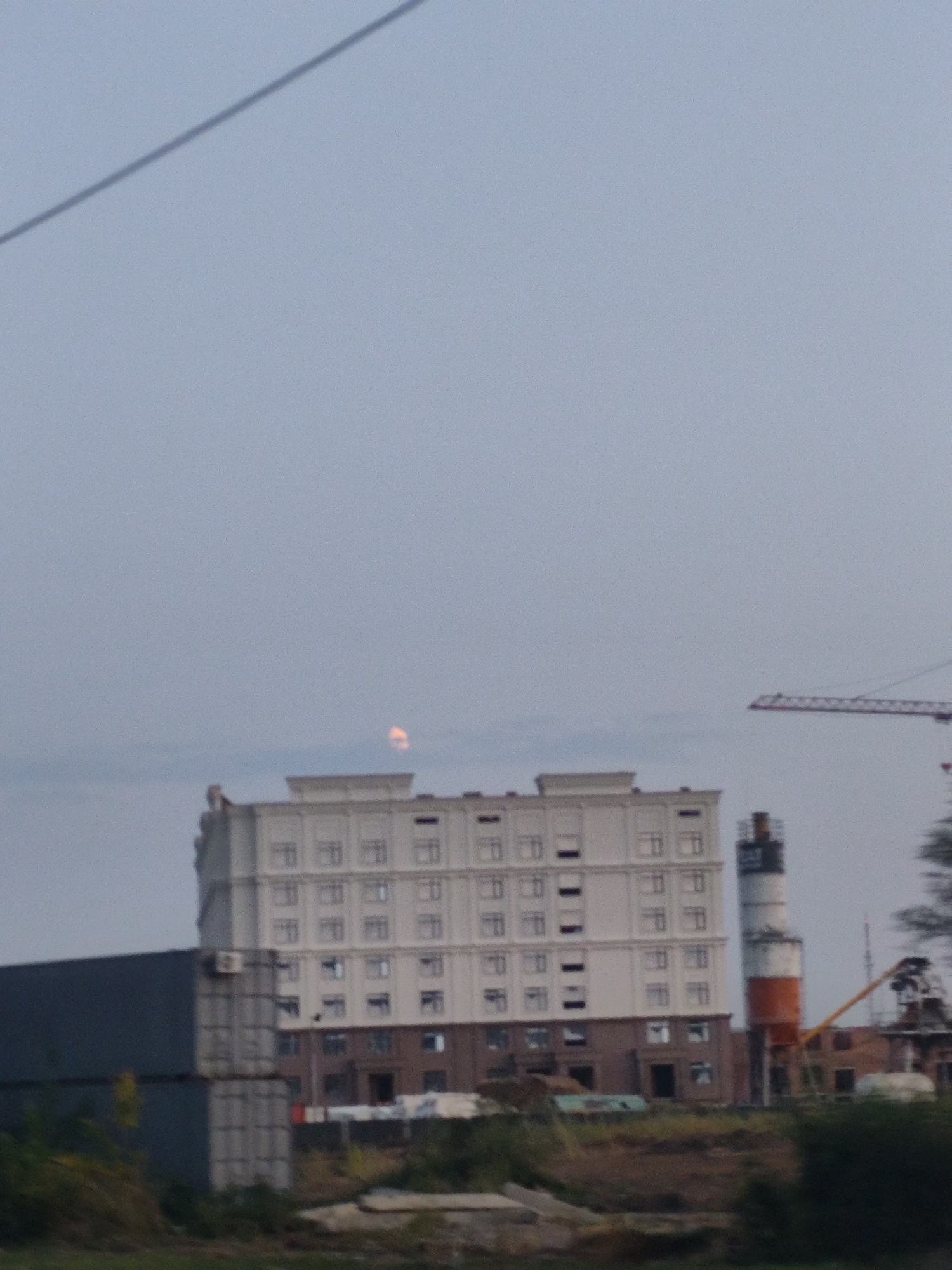 An overcast sky is visible in the background of this photograph, with a thin wire extending diagonally across the upper left corner. In the middle ground, a large, multi-story building with a light-colored facade and numerous windows stands prominently. To the right of this building, a tall, cylindrical industrial structure with alternating bands of white and orange is partially visible, and a construction crane extends from the far right of the frame. In the foreground, a dark, rectangular structure is situated to the left of the main building, with a patch of greenery and rough terrain below. The image appears to capture an urban or industrial landscape in Petropavl, Kazakhstan.