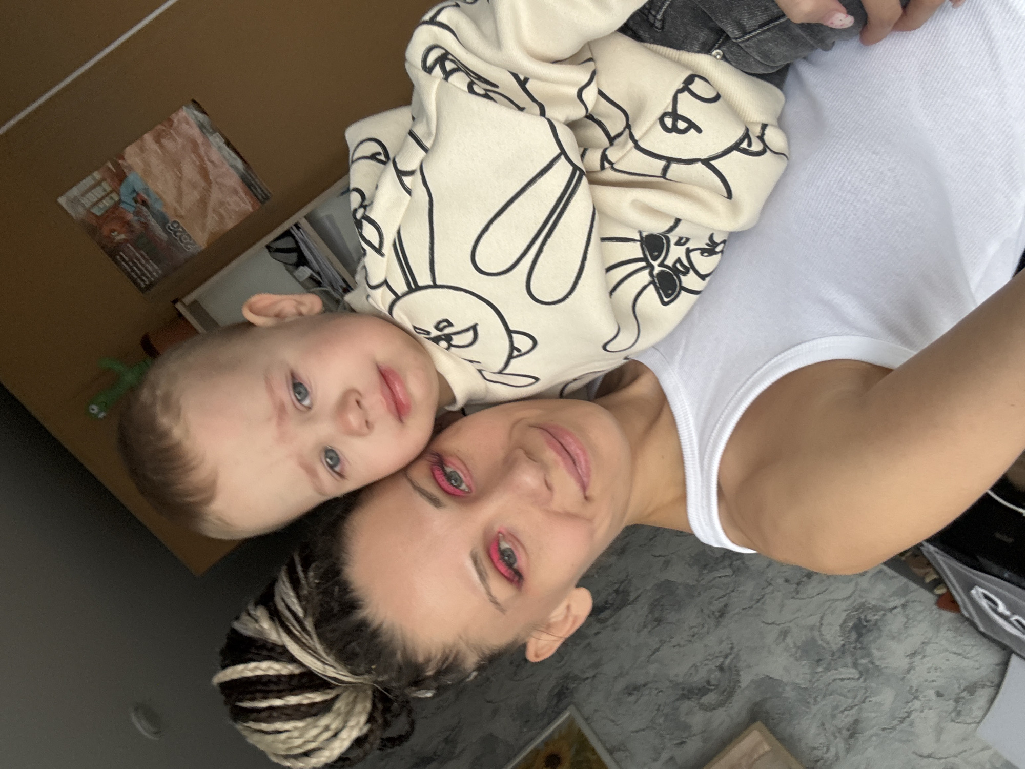A selfie shot in Rusanovo, Russia, shows a light-skinned woman with dyed blonde and brown braids in a bun, lying on her back with a young child lying on top of her. The woman has pink eyeliner and lipstick. The child has blue eyes and fair hair, wearing a cream-colored sweatshirt with a black cartoon bunny design. The woman is wearing a white tank top. The background features a box and a patterned floor.