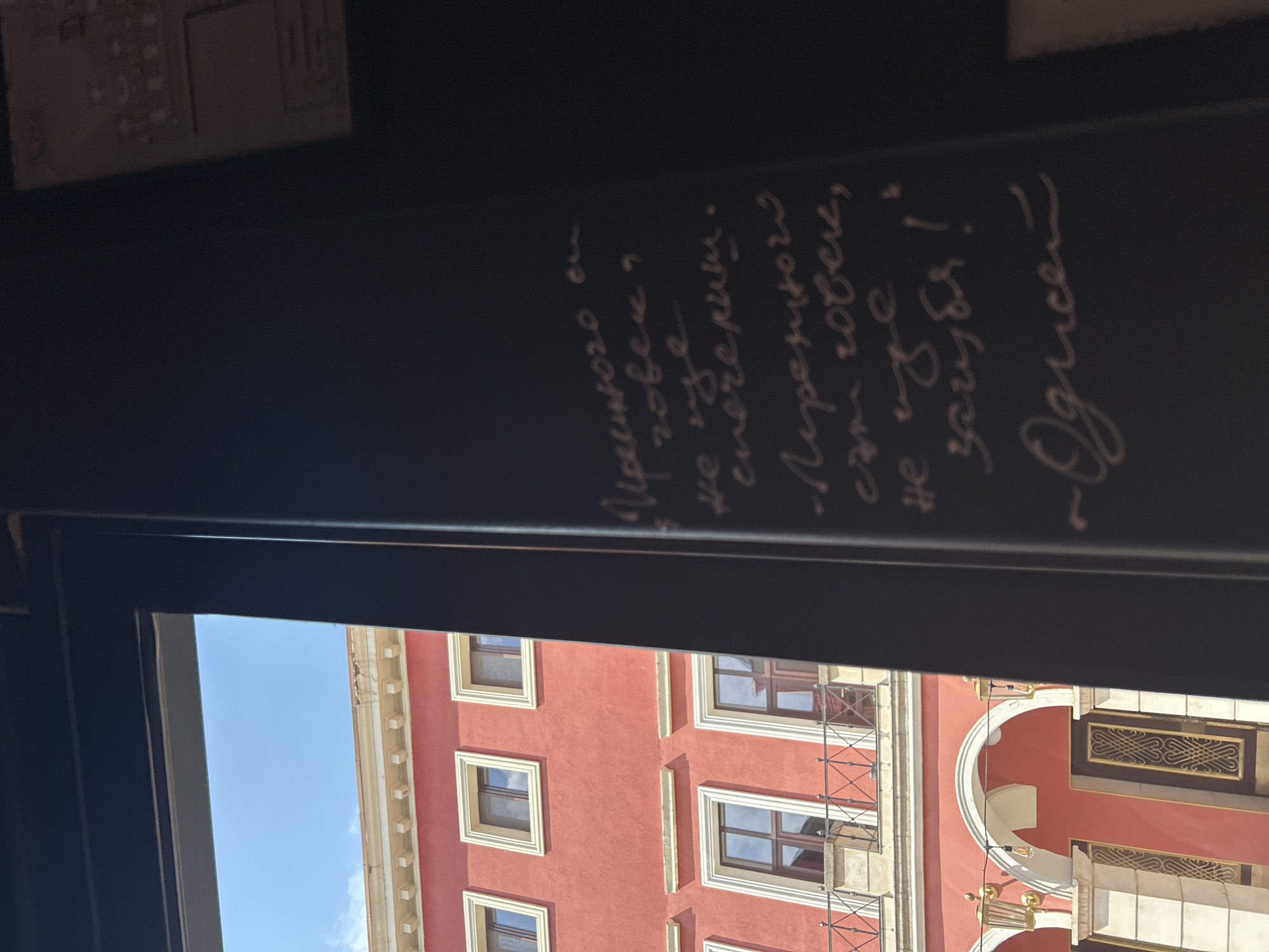 The image captures an indoor perspective looking out a window, with a prominent black surface in the foreground featuring white handwritten text. Through the window, a vibrant red building with classical architectural details is visible against a clear blue sky.
The text, written in Cyrillic script, reads: "Изреченото може да те спечели. Неизреченото те загуби! ~Одисей". This translates to: "What is spoken can win you. What is unsaid can lose you! ~Odysseus." The message conveys a thoughtful and perhaps cautionary sentiment about the importance of communication.
The red building, which dominates the outdoor view, features numerous rectangular windows framed in white or cream. It exhibits ornate cream-colored cornices and other decorative architectural elements, suggesting a historic urban environment. On the left side of the building, a structure resembling scaffolding or a decorative balcony with cross-patterned railings can be observed. To the right, part of a rounded architectural arch and a small light fixture are also visible.
The scene is set during bright daylight under a clear, cloudless blue sky, indicating sunny weather. The Cyrillic text and the architectural style are consistent with the provided context of Plovdiv, Bulgaria, suggesting a charming European city setting.