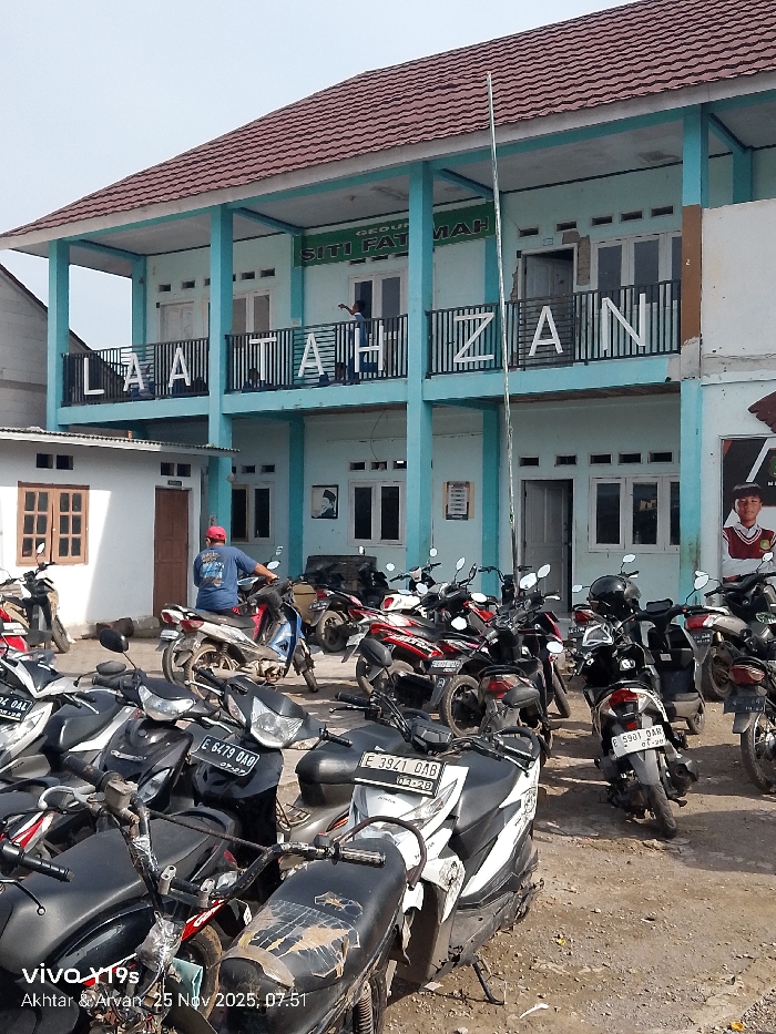 A two-story building with "SITI FATMAH" printed at the top and "LAATAHZAN" spelled out with large letters on the balcony is the focal point. A man stands on the balcony. A dense collection of motorcycles fills the foreground parking area. A small building is located to the left of the main building. A flagpole rises prominently in front of the main building. The sky is overcast. A timestamp and device watermark are visible at the bottom left.