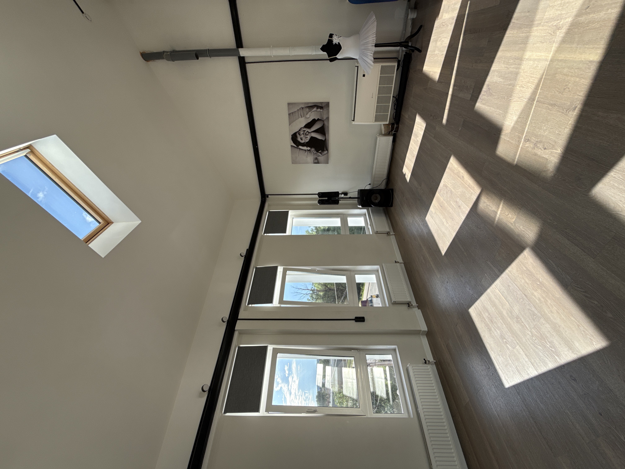 This is a wide-angle, eye-level shot of an interior space, likely a dance studio, in Zakharovo, Russia. The room features white walls and a light-colored wooden floor with distinct wood grain patterns. Natural light streams in from two large windows on the left wall, one of which is a skylight angled towards a blue sky with scattered clouds. Sunlight also casts dramatic geometric shadows across the floor, suggesting it's a sunny day, likely morning or afternoon.
The room is sparsely furnished but contains elements relevant to a studio. Along the upper part of the right wall, there's a modern white air conditioning unit mounted high, and below it, a radiator. A black and white framed photograph of a person in a ballet pose is hung on the wall. Further down, near the floor, a black speaker rests on a white radiator. A white ballet tutu on a stand is positioned near the ceiling. Several windows line the right wall, each with a view of trees and sky. Two radiators are visible beneath these windows. Metal pipes run along the ceiling. The overall impression is a clean, modern, and well-lit space. There is no visible text.