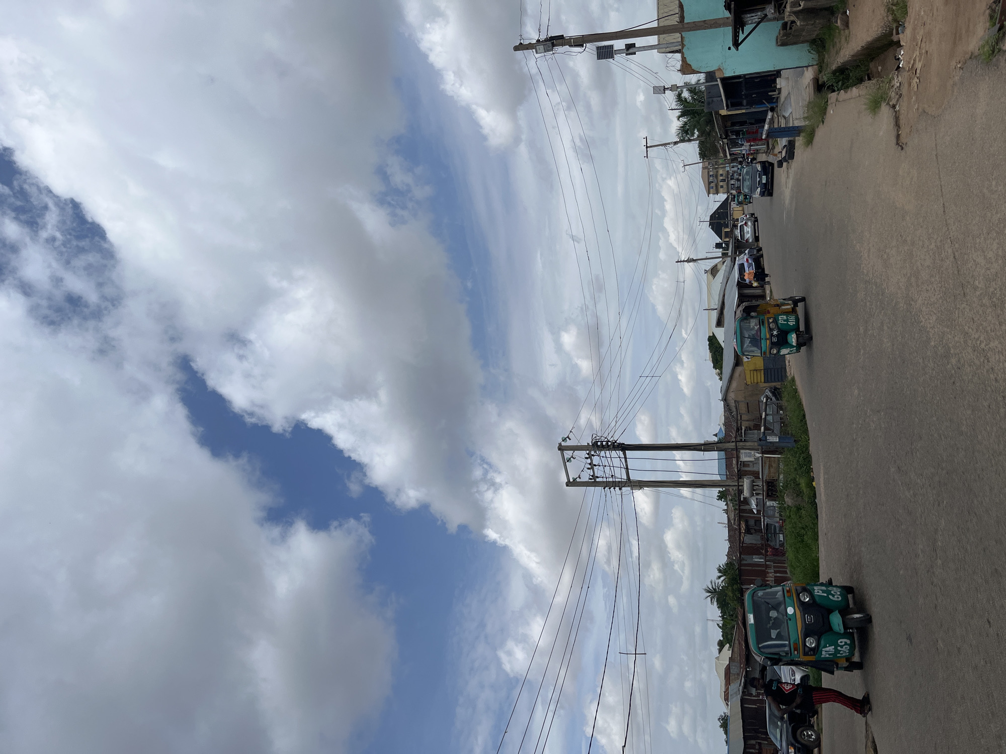 The image shows a street scene in Jos, Nigeria. The perspective is from ground level, looking up at a cloudy sky with patches of blue. A prominent feature is a utility pole with numerous wires stretching across the frame. To the right, the street is paved, with buildings lining one side. Vehicles are visible on the street, including a green and yellow auto-rickshaw with the text "PTA 6069" and "Go" visible, parked in the foreground. Behind it, several cars are parked or driving. The buildings appear to be a mix of shops and residential structures, some with corrugated metal roofs. The overall scene suggests a typical day in an urban or semi-urban environment. The weather appears overcast with some sunlight breaking through the clouds. The time of day is likely daytime, given the natural light.