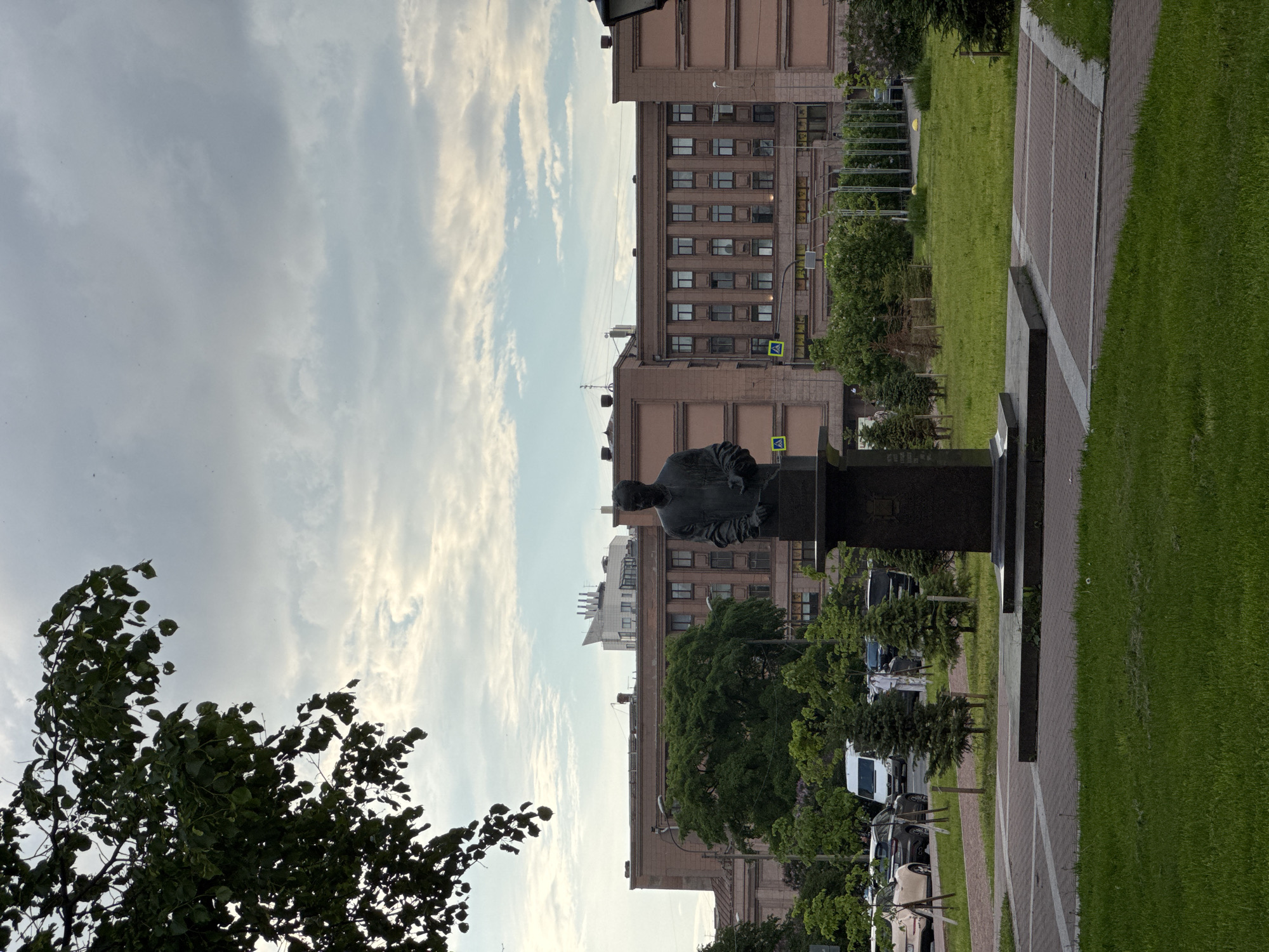 The image captures an outdoor urban scene in Saint Petersburg, Russia, centered around a commemorative statue. In the mid-ground, slightly to the right, a dark-colored bust or statue of a seated individual, possibly a man in formal wear, rests on a prominent dark stone pedestal. The statue is situated alongside a brick-paved pathway that runs horizontally through the frame, featuring lighter-colored stripes.
To the left of the statue, a verdant grassy area extends, with a large, leafy tree partially obscuring the sky in the far left foreground. To the right of the statue and pathway, more manicured green lawns lead towards a row of parked cars and vans, interspersed with smaller, conical evergreen trees.
Dominating the background are several large, multi-story buildings constructed from a reddish-brown or light brown material, characterized by numerous windows and a somewhat formal architectural style. Near these buildings, yellow diamond-shaped signs, typical of pedestrian crossings, are discernible.
The sky above is partially cloudy, displaying a mix of light gray and white clouds, indicating a daytime setting, likely late morning or afternoon, with diffused lighting. The overall scene suggests a tranquil public park or square within a city. While there appears to be engraved text on the statue's pedestal, it is not legible.