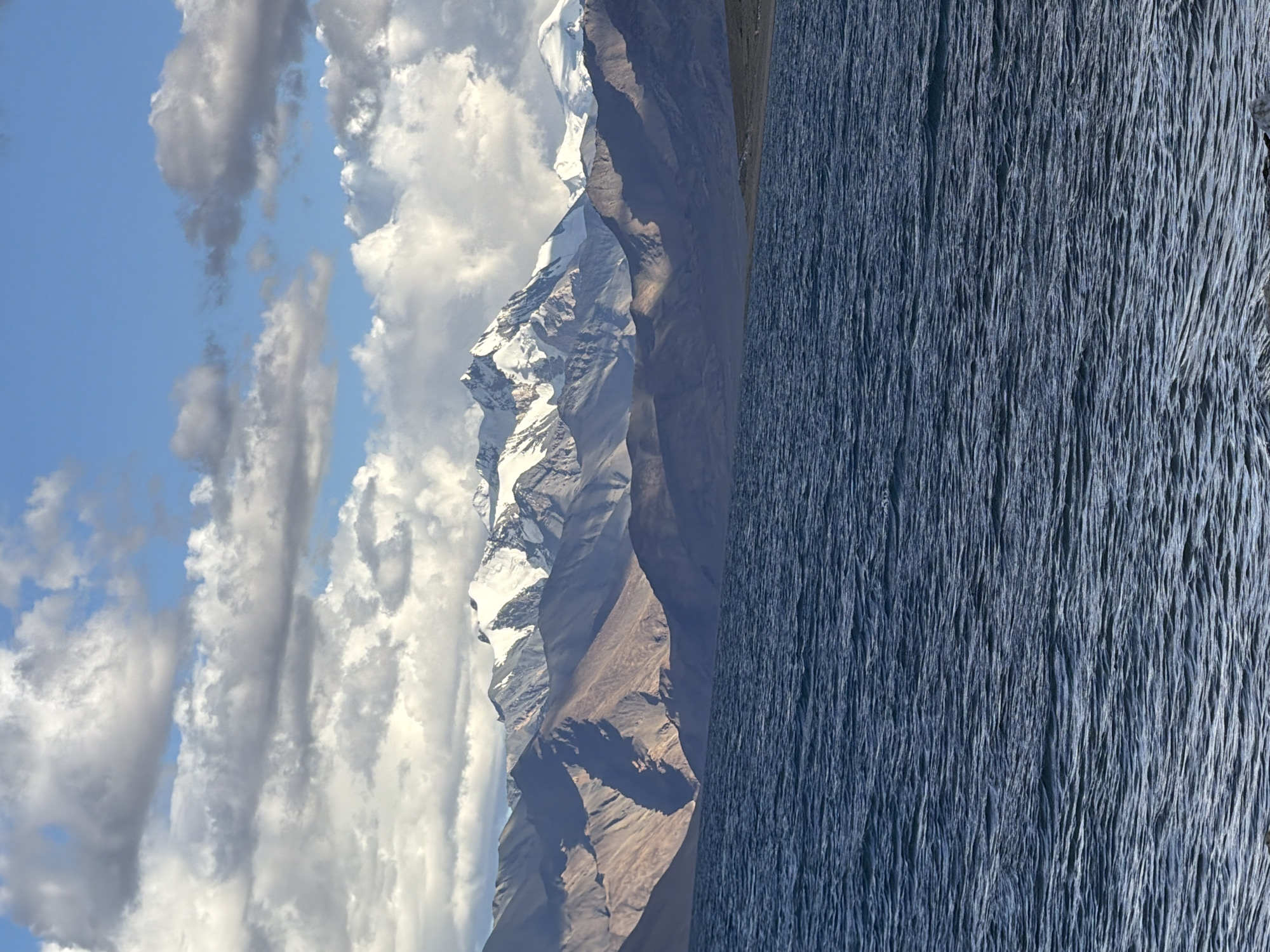 The image shows a serene lake in front of a majestic mountain range under a cloudy sky. The lake's surface is rippled, reflecting the ambient light and creating a dynamic texture. The mountains, some of which are capped with snow, rise dramatically behind the lake, their slopes exhibiting varying shades of brown and gray due to the play of light and shadow. The sky above is a mix of blue patches and scattered white and gray clouds, some of which are dense and dramatic. The scene captures a natural landscape of considerable scale and beauty, possibly evoking a sense of tranquility and awe. The location is identified as the city of Karzok, India.
