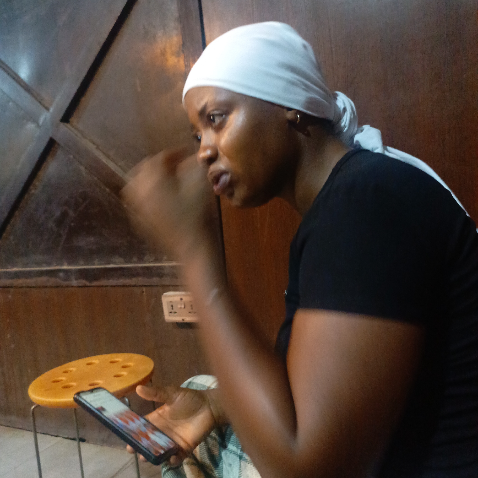 The image depicts a woman in an indoor setting, possibly in the city of Unknown, Unknown. She is seated, wearing a white headscarf and a black shirt. She is holding a smartphone in her left hand and gesturing with her right. Beside her is a yellow stool with holes in the seat. Behind her is a wooden structure, possibly a door or a wall panel, and an electrical outlet is visible. The lighting appears to be somewhat dim and the overall environment seems simple and informal.