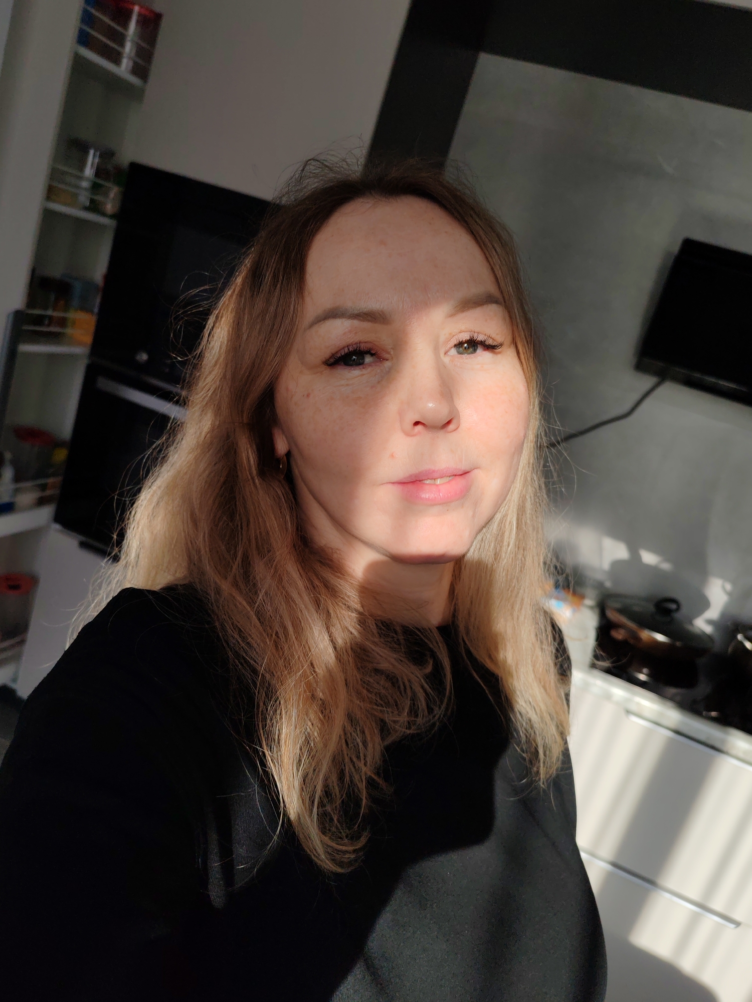 A woman with fair skin, freckles, and blonde hair poses for a selfie indoors. She is wearing a black top. Sunlight illuminates part of her face. In the background is a kitchen with a black oven, microwave, white cabinets, and a television on the wall. The location is Chaikovsky, Russia.
