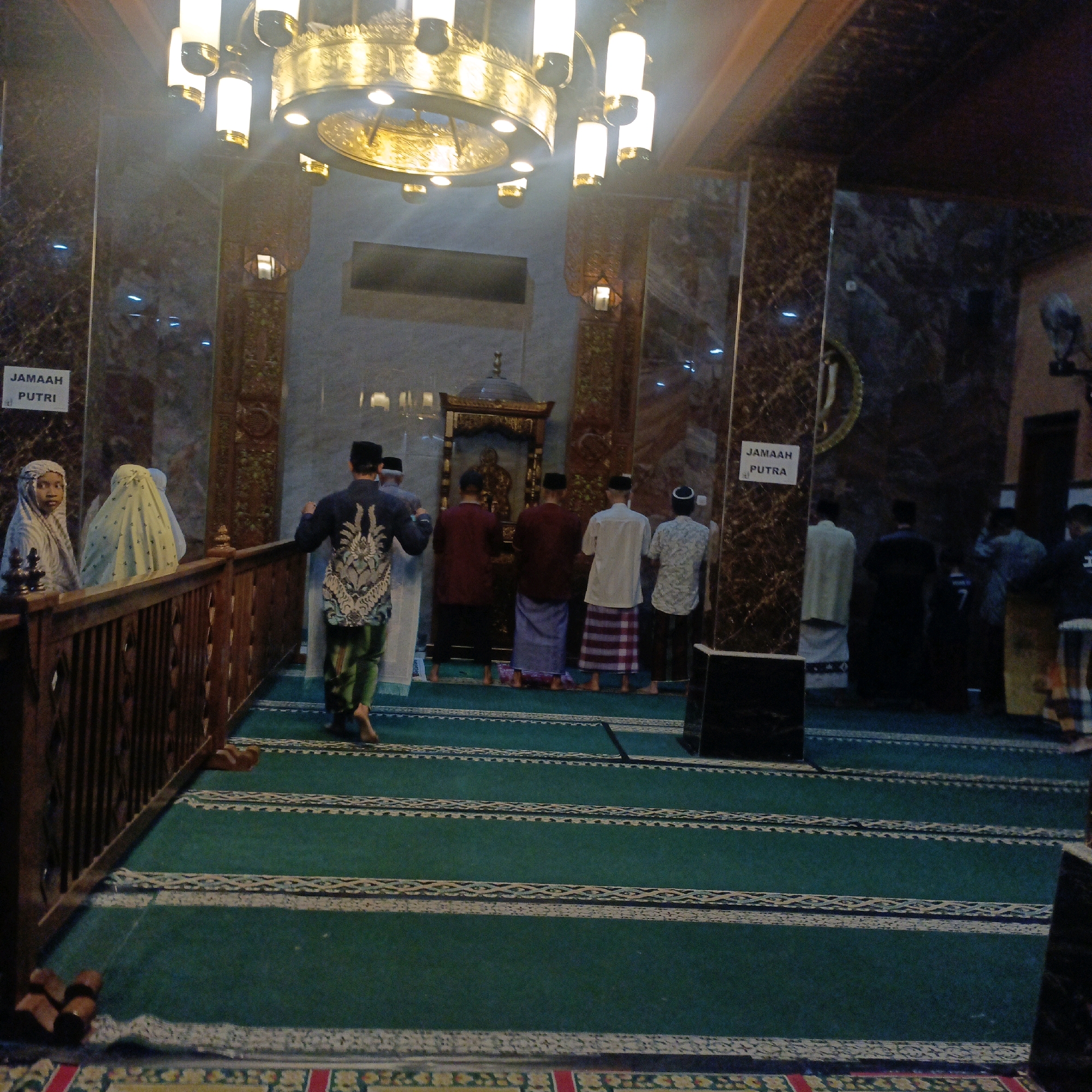 The image depicts the interior of a mosque, likely in Salatiga, Indonesia, during prayer time. Several men are seen standing in prayer rows facing a mihrab, an ornamental niche in a mosque that indicates the qibla, the direction of Kaaba in Mecca. Some individuals are wearing traditional Muslim attire, including caps and sarongs. On the left side of the image, there is a section partitioned by a wooden railing, where two women wearing hijabs are present. Signs are visible indicating separate prayer areas for "JUMAAH PUTRI" (Women's Congregation) and "JUMAAH PUTRA" (Men's Congregation). A large, ornate chandelier hangs from the ceiling, illuminating the space. The floor is covered with green prayer mats featuring intricate patterns. The mosque interior features marble-patterned columns and walls, contributing to a grand and solemn atmosphere. The lighting appears to be a mix of natural light and artificial illumination from the chandelier.