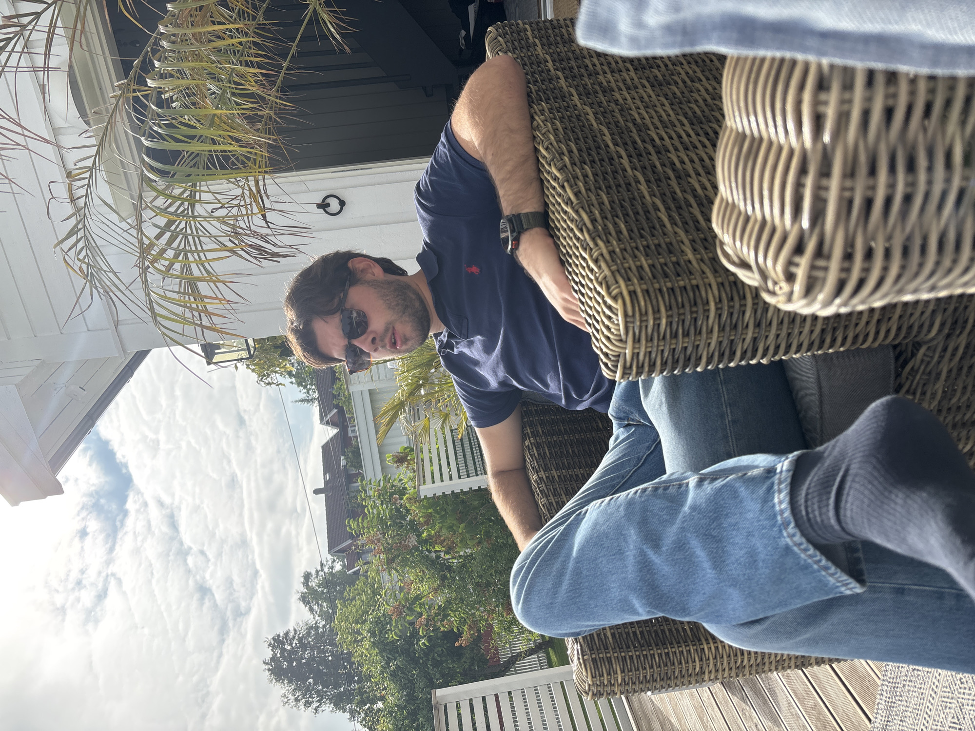 This image depicts two individuals relaxing outdoors on a sunny day. The foreground features a man wearing a navy blue polo shirt, blue jeans, sunglasses, and a watch. He is seated in a woven wicker chair, his legs crossed. The second person is partially visible, with their legs also crossed and clad in blue jeans, seated in a similar wicker chair. A light blue fabric drapes over the back of one of the chairs.
The setting appears to be a patio or deck, with weathered wooden planks visible beneath the chairs. A white railing runs along the edge of the patio, and beyond that, lush green trees and houses with reddish-brown roofs are visible. The sky is bright blue with scattered white clouds, suggesting pleasant weather and a mid-day time frame. A palm-like plant with fronds droops into the frame from the upper left, adding a touch of tropical atmosphere to the scene. The overall mood is relaxed and casual. No visible text is present in the image. The location context provided is Larkollen, Norway.