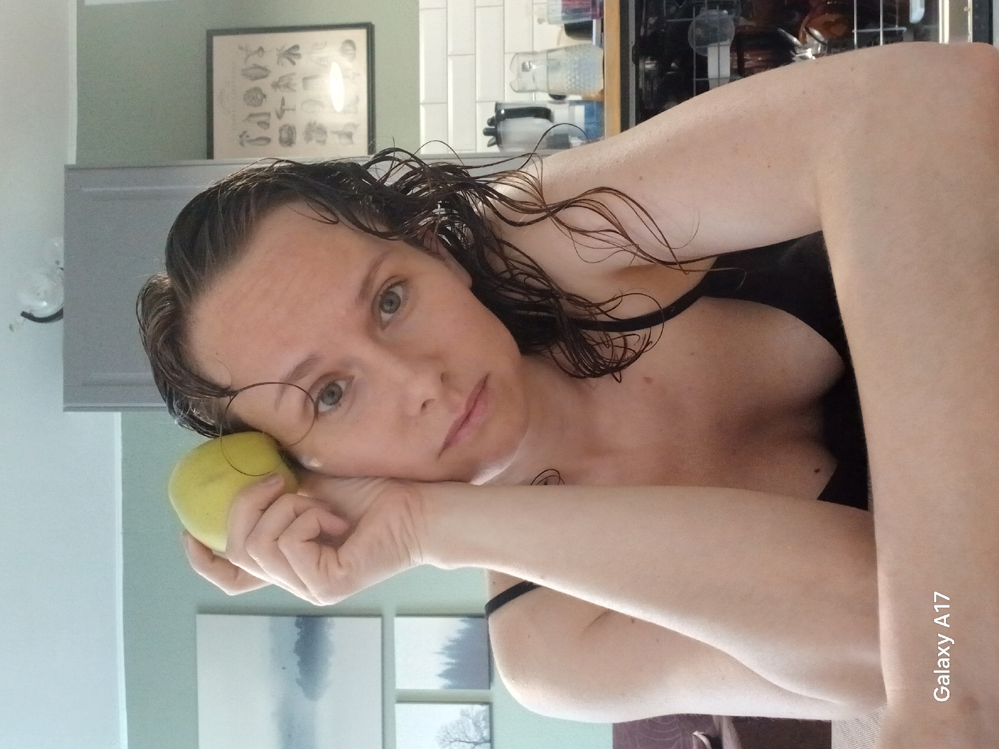 A woman with wet, dark hair is holding a green apple near her face, as if resting her cheek on it. She is wearing a black tank top. The background includes a kitchen area with white subway tiles, a framed botanical print, a light gray cabinet, a dishwasher, and what appear to be decorative bottles or dispensers. Below the cabinet are three framed pictures with landscapes. The camera watermark "Galaxy A17" is visible in the bottom right corner. The photo is rotated. There's no evidence to indicate this is in Yekaterinburg, Russia.