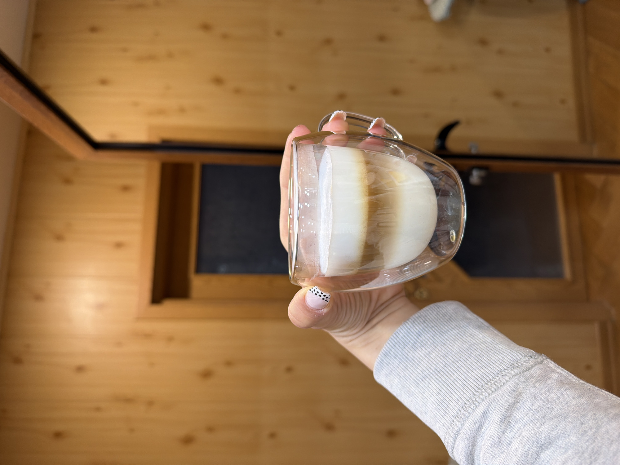 A hand in a grey sweatshirt sleeve holds a clear, double-walled mug containing a layered beverage of brown liquid and white foam in Buhovo, Bulgaria. The hand's nails have a light color with patterned tips. The background is a light brown, wood-paneled wall with a dark framed window.