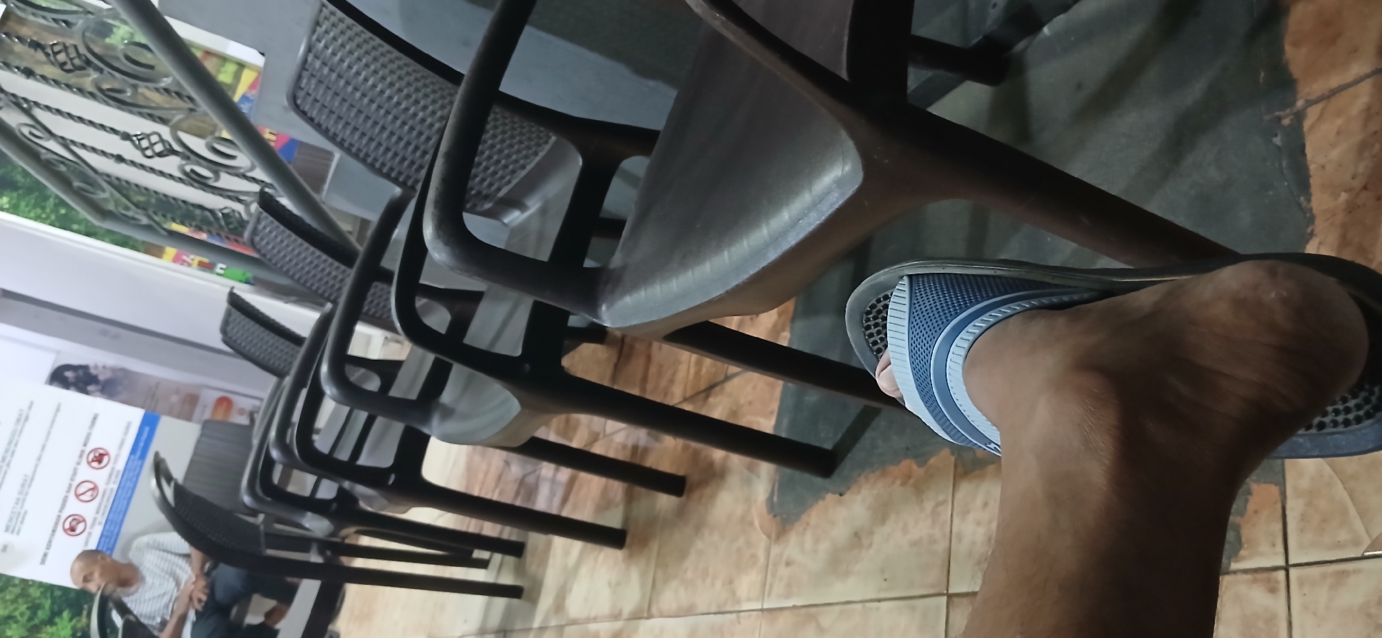 The image depicts a waiting area with multiple rows of dark plastic chairs. In the foreground, a person's left foot wearing a blue and white sandal is visible. The floor is tiled with a pattern that appears worn in places. In the background, a man is seated in one of the chairs, looking to his left. A sign with text and graphics is mounted on the wall behind the man. The scene suggests a public space, possibly a government office or clinic, given the presence of the waiting chairs and informational sign. The overall lighting is dim, with some light coming from the right side of the frame. This location is identified as being in Samarinda, Indonesia.