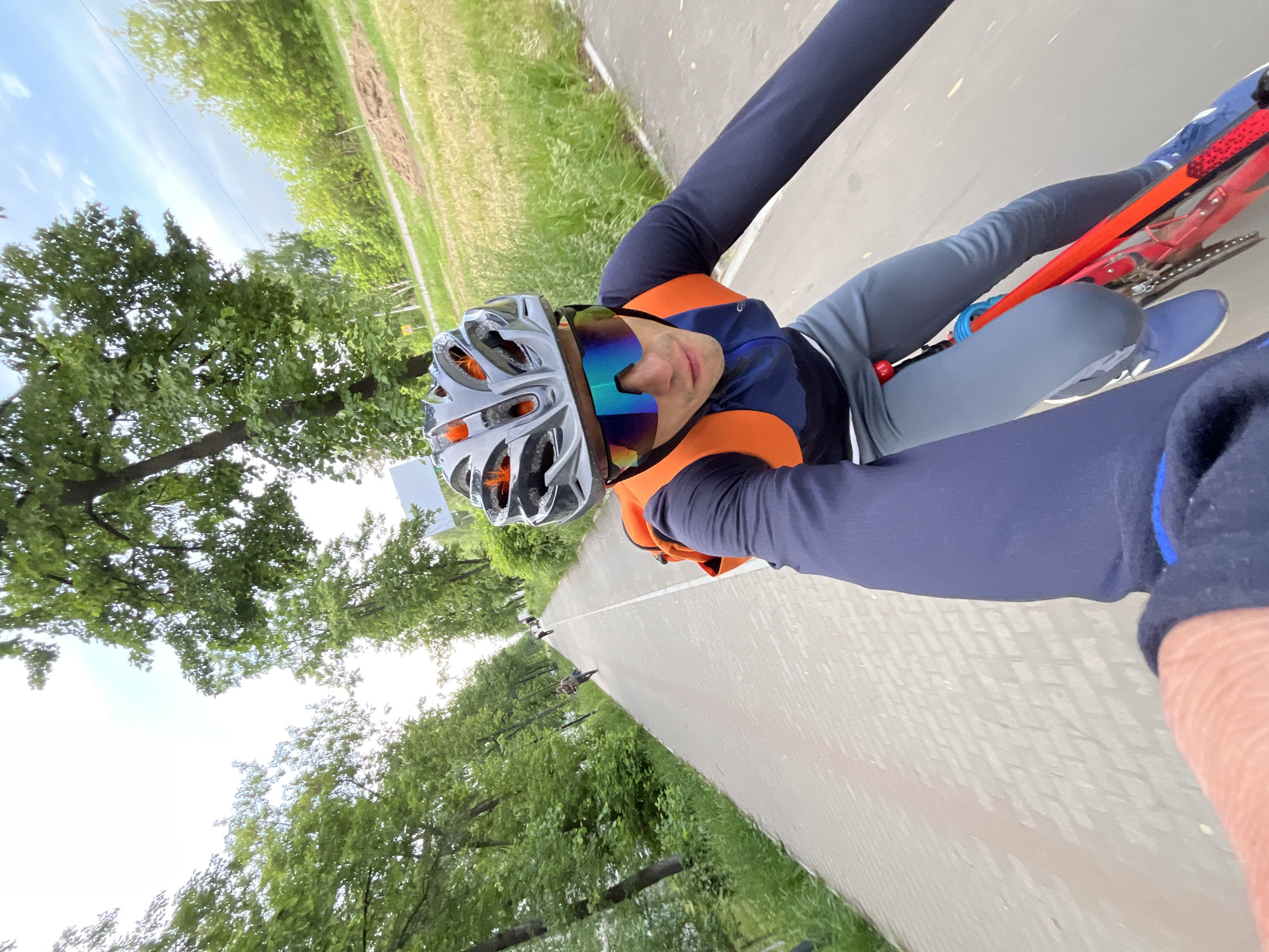 A cyclist is taking a selfie on a paved path lined with trees and grass. The path appears to be located in the city of Chaikovsky, Russia. The cyclist wears a helmet and sunglasses and is dressed in cycling apparel. The bicycle is partially visible. Other cyclists can be seen further down the path.
