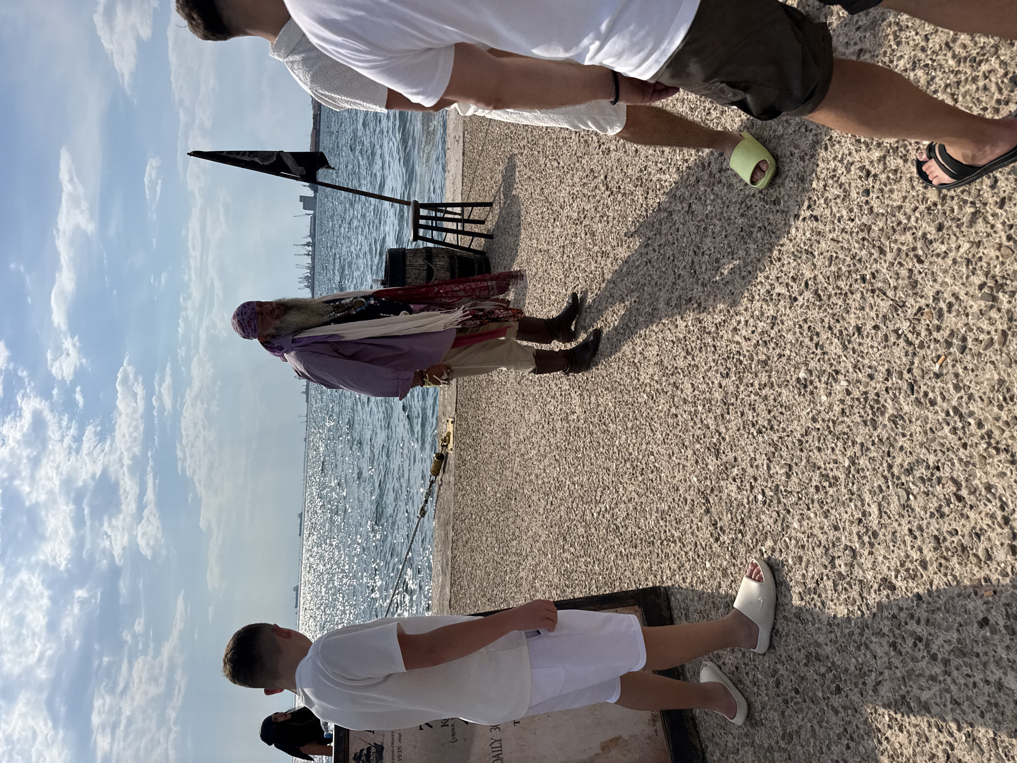The image depicts an outdoor scene along a waterfront promenade, likely in the city of Thessaloniki Municipal Unit, Greece, given the presence of a pirate-themed entertainer. In the foreground, a young boy in a white t-shirt and white shorts stands with his back to the camera, facing towards the sea. To his left, a portion of a signpost is visible.
To the right of the boy, a man dressed as a pirate stands near a barrel and a stool. He has a long white beard, a purple bandana, and is wearing a light purple open shirt over a dark vest adorned with jewelry. He also wears light-colored pants tucked into brown boots. Several colorful scarves hang from his neck.
Further to the right, two other individuals are partially visible, facing away from the camera. One wears a white patterned shirt and dark shorts, while the other wears a white t-shirt. A black pirate flag is visible behind the pirate character, attached to a pole.
The background features the shimmering blue sea under a partly cloudy sky. In the distance, industrial structures and cranes are visible along the coastline. The ground is covered with a rough, pebbly surface. The overall impression is that of a public space possibly during a festival or tourist attraction.