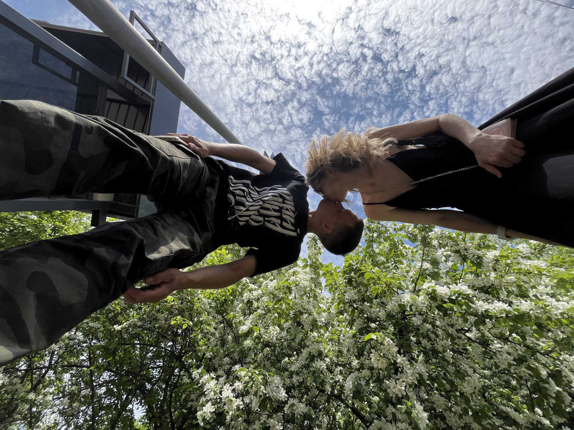 A low-angle shot captures a man and a woman kissing, with their bodies at an angle relative to the camera. The man is on the left, wearing camouflage pants and a black T-shirt with a white graphic design. His hand rests on a metal railing. The woman, on the right, wears a black dress with thin straps and holds a small clutch purse.
The background is complex. A blue sky filled with patchy, white clouds is visible above. Below the couple, a tree with white blossoms is in full bloom. Architectural elements, including a metal structure and a partially visible building, are present in the top-left corner.
The photo was taken in the city of Chaikovsky, Russia.