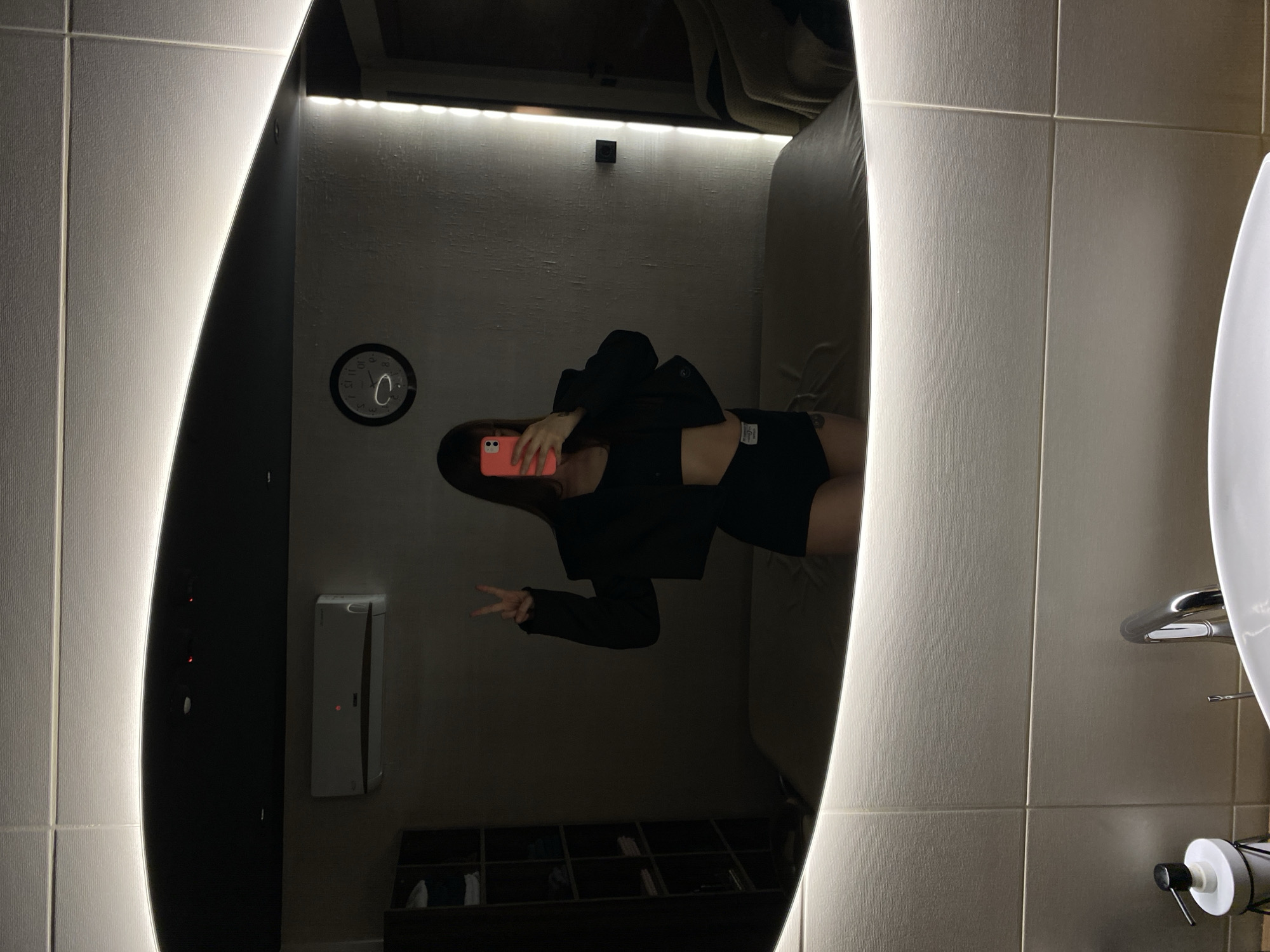 The image displays a selfie taken in a large, vertically oriented mirror within an indoor setting, likely a bathroom or dressing room. The mirror features integrated LED strip lighting along its vertical edges, providing illumination.
The main subject is a person, visible in the mirror's reflection, taking the selfie with a light pink or coral-colored smartphone held in front of their face. They are dressed in a black cropped top, black shorts, and an open black long-sleeved jacket. The person's left hand is raised, making a peace sign gesture. A tattoo is visible on their upper thigh/hip area in the reflection.
Behind the person, as seen in the reflection, is a dark wall. A round black clock with white numbers (such as 12, 3, 6, 9) is mounted on this wall, showing the time to be approximately 11:55 or 12:55. Above the clock, a light strip illuminates the textured wall. To the right of the clock, a small, white rectangular object, possibly a hand dryer or air freshener, is mounted on the wall. In the upper part of the reflected room, a light-colored, soft surface, resembling a sofa or bed, is partially visible. Below the person's reflection, open shelves containing folded items are visible.
Outside the mirror's frame, to the right, is a tiled wall with a partial view of a white sink, a silver faucet, and a white soap dispenser. The wall tiles have a subtle texture. The overall ambiance is dim, with the mirror's integrated lights serving as a prominent light source. The only clearly visible text is the numerals on the clock face.