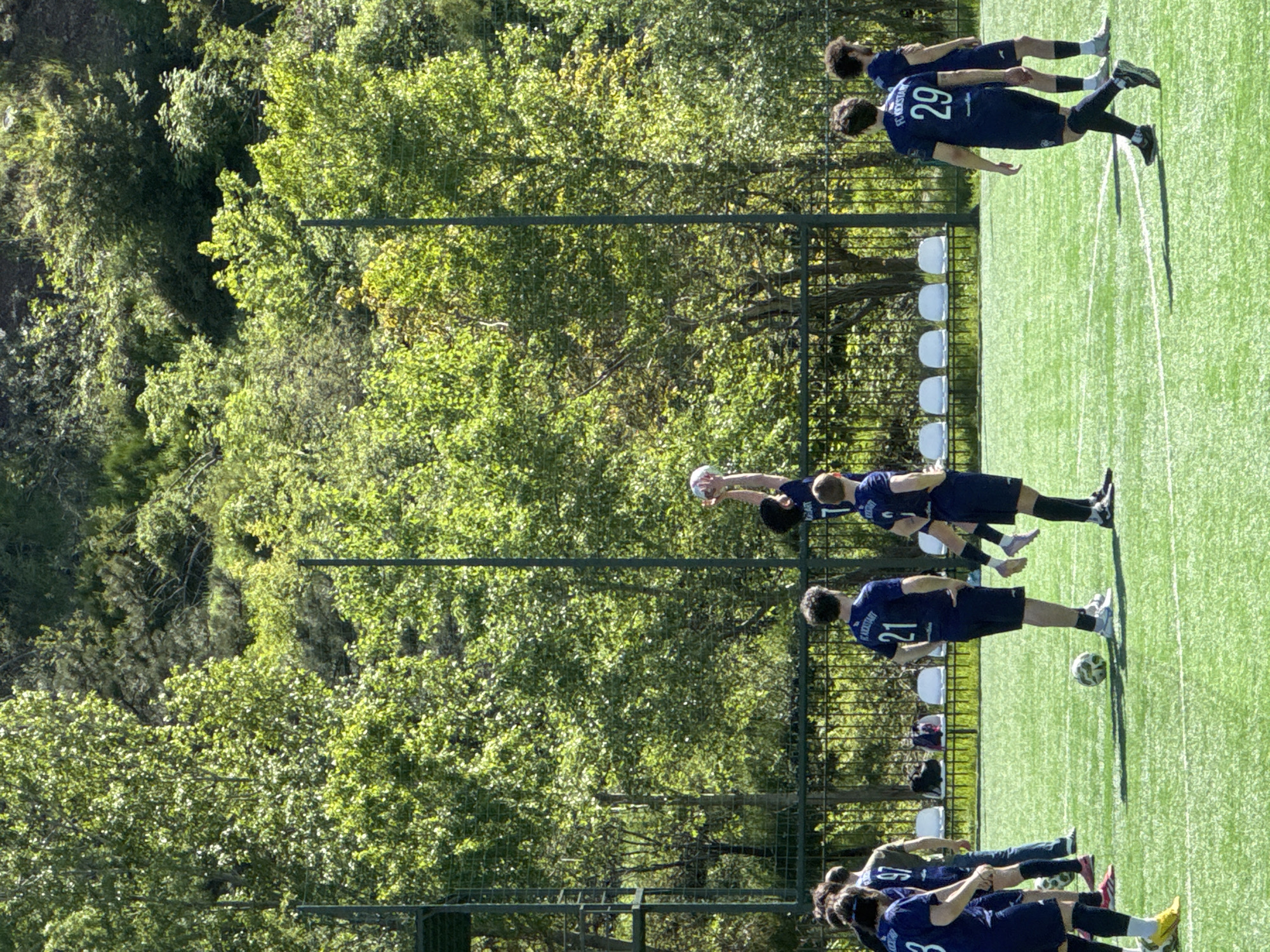 A medium-shot photograph shows a soccer practice on a green field surrounded by trees. A metal fence separates the field from the trees. Four soccer players in matching blue uniforms are in a line facing right, with the player furthest from the camera juggling a soccer ball with their head. Behind them are six white chairs. A player in a blue uniform faces right with a soccer ball at their feet. Further to the bottom of the image, three soccer players in blue uniforms face left, with one touching a soccer ball with their foot. The team's uniform has "FC KICKSTART" printed on the back. The location appears to be the city of Tbilisi, Georgia.