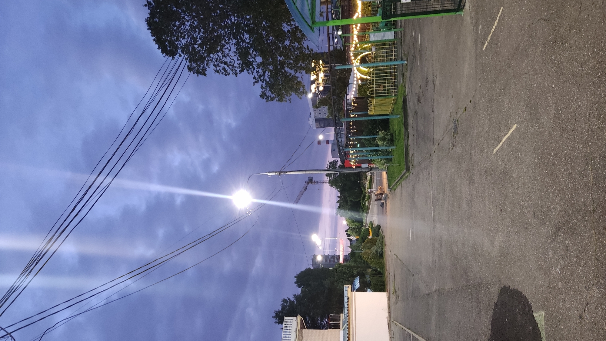 The image depicts an outdoor scene at dusk or dawn, with a cloudy sky tinged with purple hues. A paved road or pathway leads into the distance, flanked by trees and some low-lying structures. Streetlights are illuminated, casting bright beams of light. In the background, a construction crane is visible, along with several buildings, one of which appears to be a multi-story modern structure. The right side of the image shows a fenced-off area with decorative lights, suggesting a park or recreational space. The overall impression is of an urban or suburban environment transitioning from daylight to night. The presence of the crane suggests ongoing construction or development in the area. There are no people visible in the image. The scene is likely located in Krasnodar, Russia, given the provided context.
