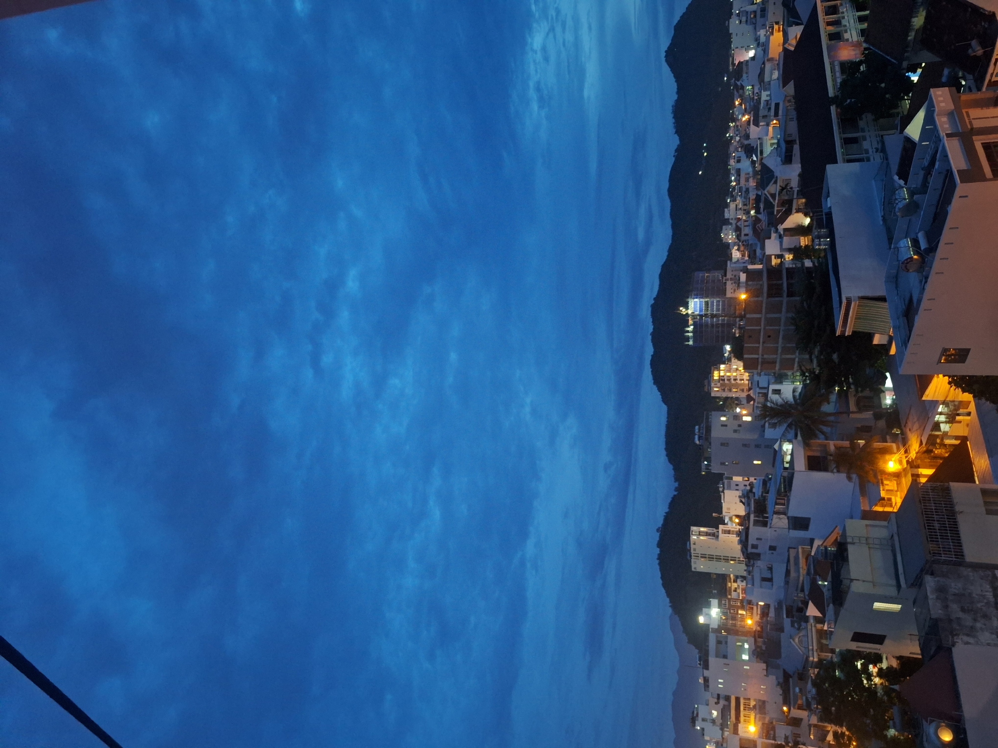 A city skyline at dusk in Nha Trang, Vietnam, is depicted. The sky is a deep, vibrant blue with wispy clouds, suggesting the transition from daylight to night. A dark, diagonal line, likely a power line, cuts across the upper left corner of the image. Below the sky, the cityscape unfolds with numerous buildings of varying heights, predominantly white or light-colored, interspersed with darker structures and some under construction. Many windows are illuminated with warm, yellow light, indicating that people are inside and the city is coming alive with evening activity. The buildings are tightly packed together, forming a dense urban landscape. In the background, silhouetted against the twilight sky, are rolling green hills or mountains, characteristic of Nha Trang's geography. The overall impression is one of a bustling coastal city at the end of the day, with a clear view of the sky and its natural surroundings.