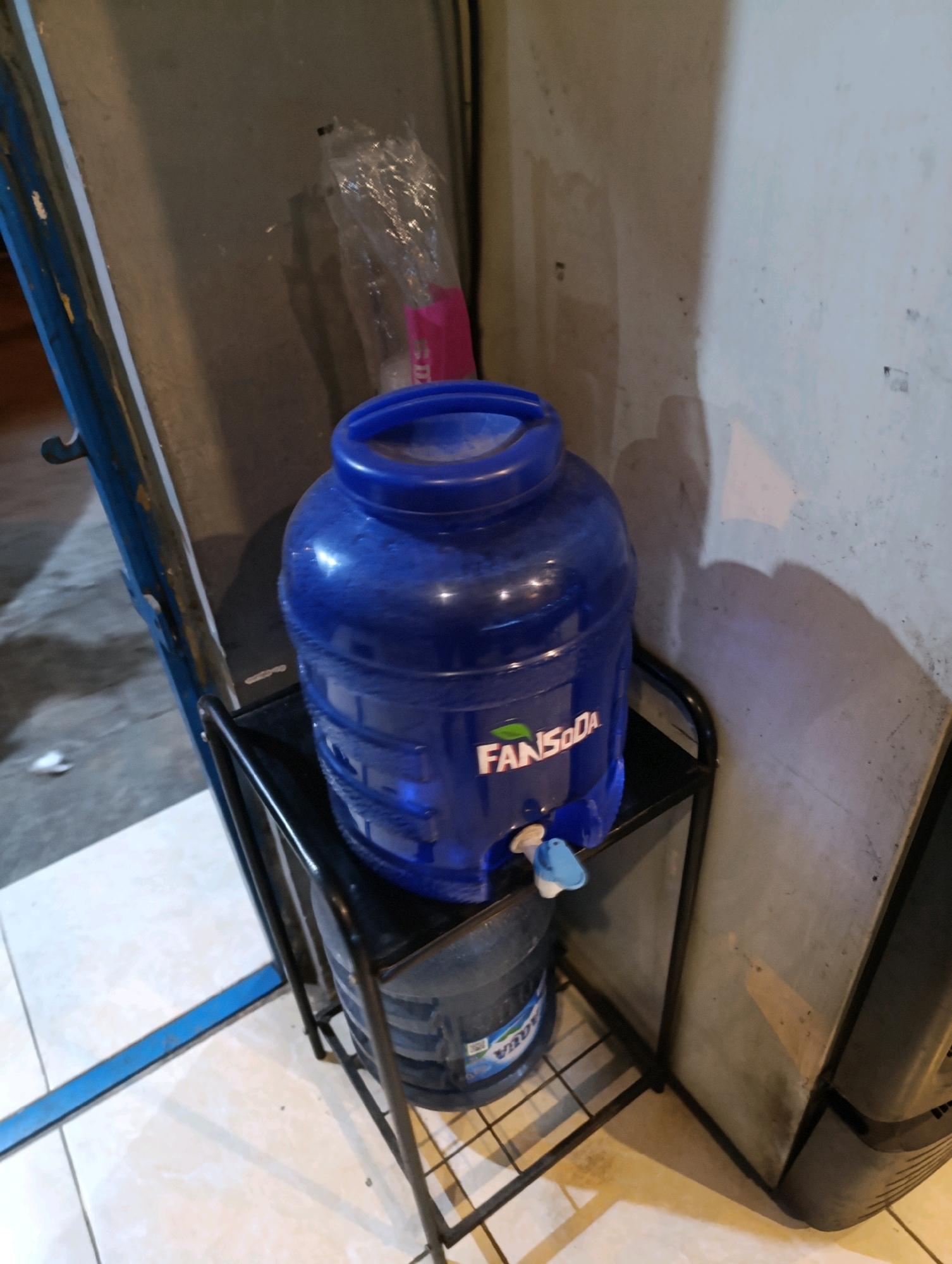 In Karangsono, Indonesia, a blue plastic water jug with the "FAN Soda" logo sits on a black metal rack. The jug has a spigot at the bottom. Beneath it, another water jug, likely larger and transparent, is visible. The rack is placed on a tiled floor, adjacent to a textured wall. A portion of a blue metal structure is also visible to the left of the scene.