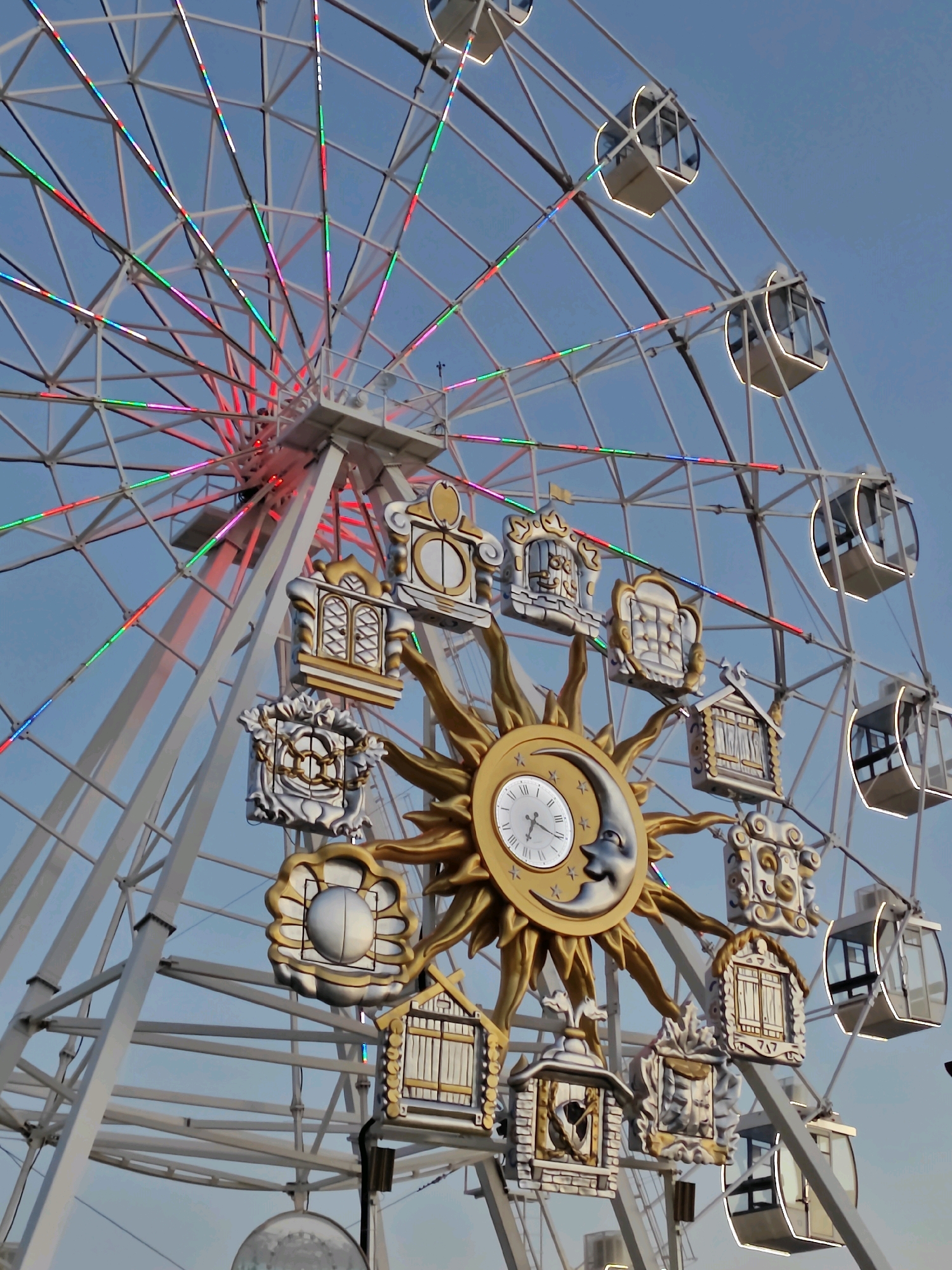 The image depicts a large Ferris wheel, decorated with numerous whimsical elements, set against a clear blue sky. The Ferris wheel has a white metal structure with a series of gondolas suspended from its outer rim. The gondolas are enclosed cabins with windows, some of which are outlined in a soft, warm light. Along the spokes and rim of the Ferris wheel, there are strings of colorful LED lights, emitting hues of red, pink, green, and blue, suggesting it is either dusk or nighttime, or the lights are on during the day for decorative purposes.
Prominently featured in the center of the Ferris wheel is a large, ornate golden sun decoration. Within the sun, there is a clock face with Roman numerals and clock hands, indicating the time. Next to the clock, a stylized crescent moon with a face is depicted. Surrounding the central sun and clock are numerous smaller, intricately designed decorative elements, resembling miniature houses and architectural structures, also in gold and silver tones, with various textures and intricate detailing. These decorative elements appear to be themed, possibly relating to fairy tales or folk art.
The overall impression is one of a festive amusement park or recreational area. Based on external knowledge, this Ferris wheel is likely located in the city of Listvyanka, Russia, a settlement on the southwestern shore of Lake Baikal. The environment appears clean and well-maintained. No people are clearly visible within the gondolas or on the ground near the Ferris wheel. The focus is entirely on the architecture and decoration of the amusement ride itself.