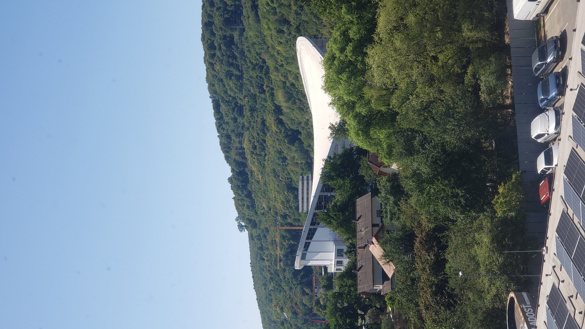 The image shows a panoramic view of a city area with a large, modern building featuring a curved white roof in the mid-ground. This building is situated against a backdrop of a lush, tree-covered hill under a clear blue sky. In the foreground, there are houses with tiled roofs and a section of a building with solar panels. Several cars are parked in a lot behind a grey metal fence. The context provided indicates this scene is located in Prešov, Slovakia, a city known for its historical architecture and natural surroundings, consistent with the image's depiction of urban structures and a significant natural landscape. The overall impression is one of a bright, sunny day with a mix of contemporary architecture and verdant nature.