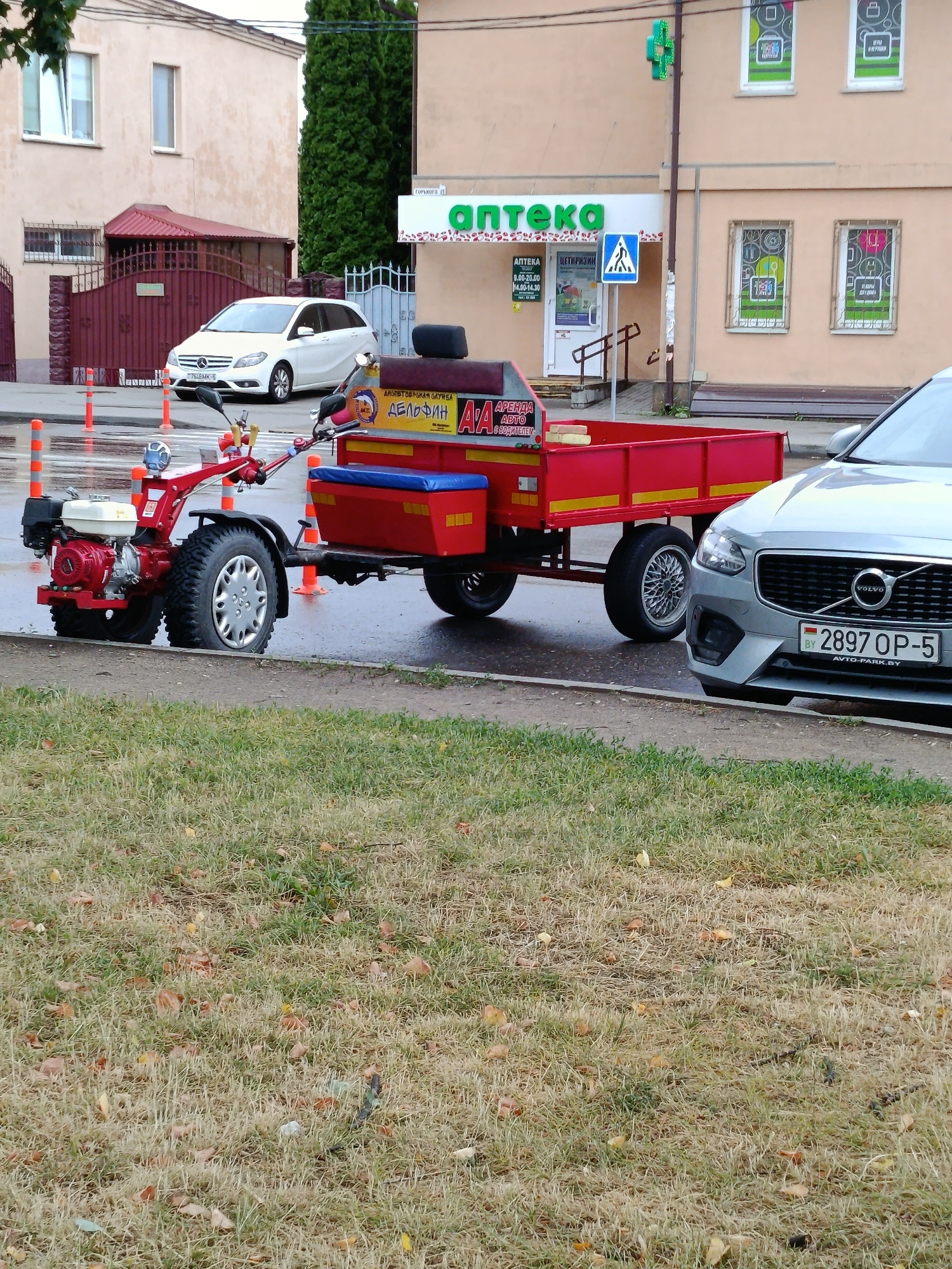A red tractor-trailer combination with a single wheel on the trailer is parked on a wet asphalt surface next to a grassy area. The tractor has a red frame and black tires, with a white engine. The trailer is red with yellow trim and a blue seat. Signs on the trailer indicate "АРЕНДА АВТО С ВОДИТЕЛЕМ" and "ДЕЛФИН".
In the background, a silver Volvo XC90 is parked, facing right, with a Belarusian license plate reading "2897 OP-5". Further back, a white Mercedes-Benz hatchback is parked in front of a building with a red roof and a dark metal fence. A pharmacy with a green cross logo and the word "АПТЕКА" is visible on the building to the right. Orange traffic cones are scattered along the roadside. The environment appears to be an urban or suburban street, possibly in the village of Smalyavitsky Selsovet, Belarus, judging by the license plate format. The ground is wet, suggesting recent rain. The grass in the foreground is dry and scattered with fallen leaves.