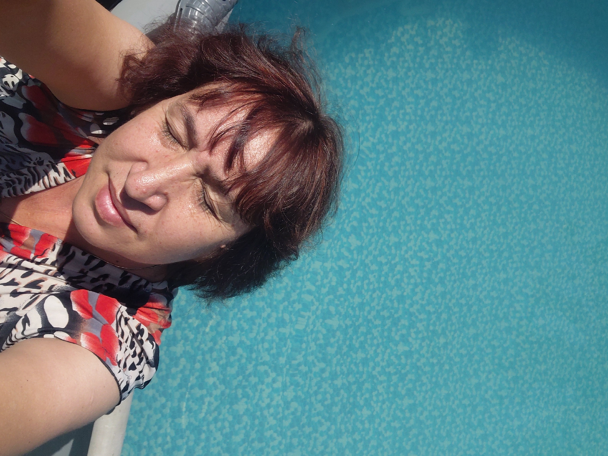 A woman with shoulder-length, auburn hair is pictured from a high angle, looking down with her eyes closed. She is resting her head on her left arm and has a faint smile on her face. The woman is wearing a colorful, patterned blouse with red, white, and black designs. She is positioned in front of a bright blue swimming pool, which occupies the majority of the frame. The water has a textured, speckled pattern. A portion of the pool's edge, a white railing, is visible in the lower right corner of the image.