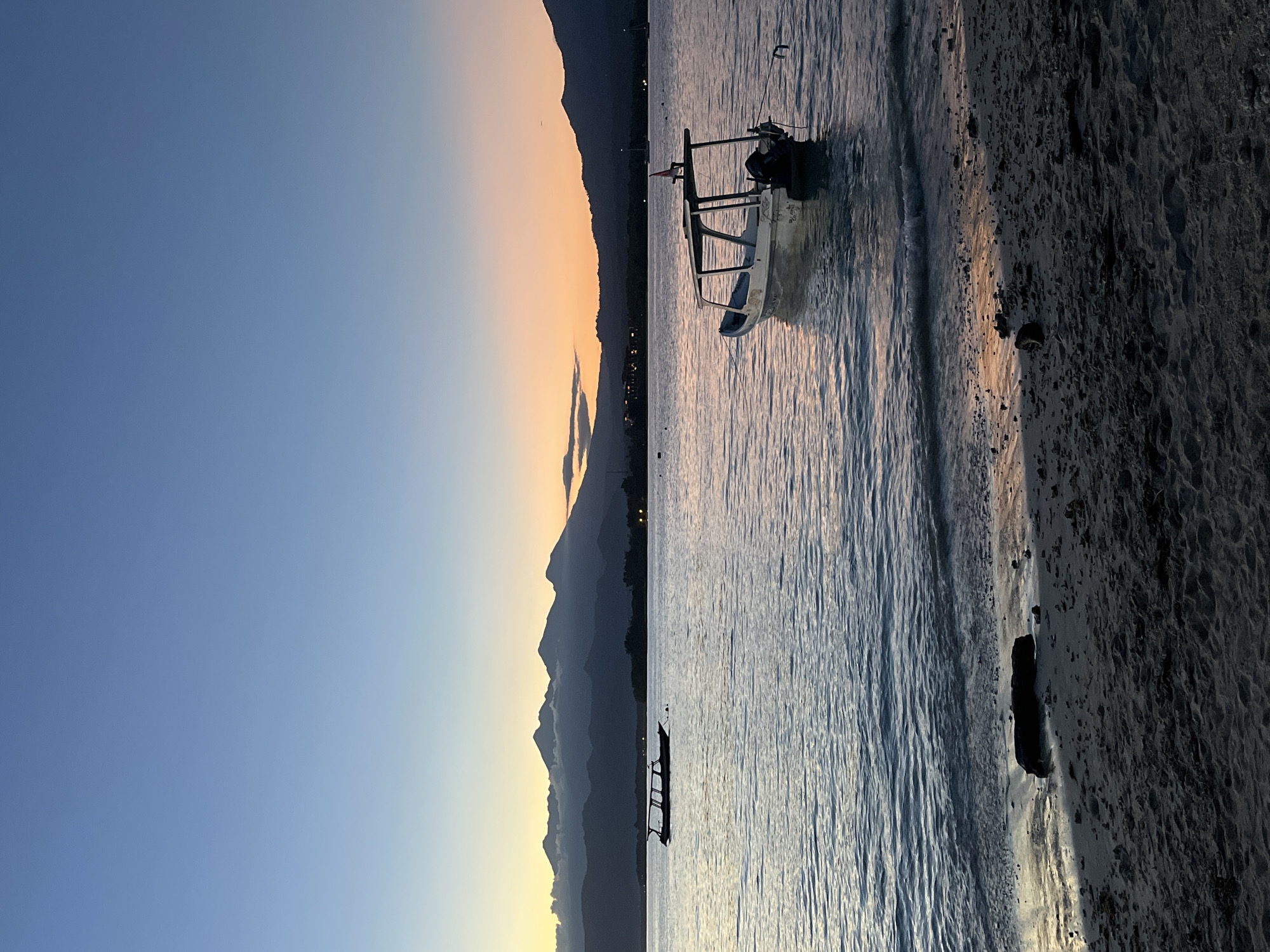 The image shows a beach scene at dawn in Unknown, Indonesia. The sky transitions from deep blue to a pale orange near the horizon, where silhouetted mountains are visible. Two small boats are in the water near the shore, and a larger boat is closer to the beach. The beach consists of dark sand and water ripples along the shoreline.
