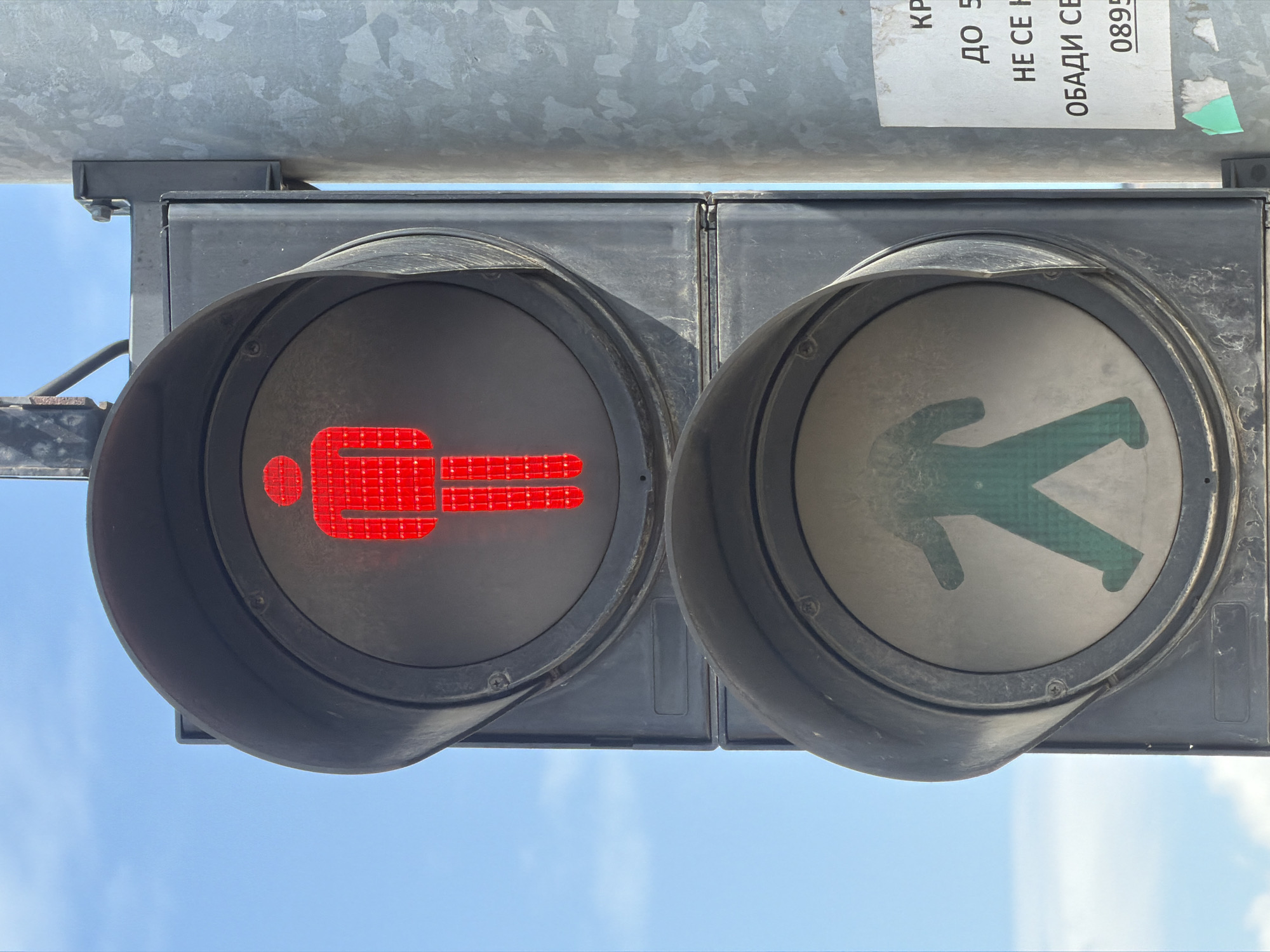 The image shows a pedestrian traffic light in Sofia, Bulgaria. The left light displays a solid red figure indicating "Do Not Walk", while the right light displays a green walking figure indicating "Walk". The traffic light is mounted on a metal pole. A sticker with Bulgarian text, including a phone number, is attached to the pole above the light. The background is a blue sky with light clouds.