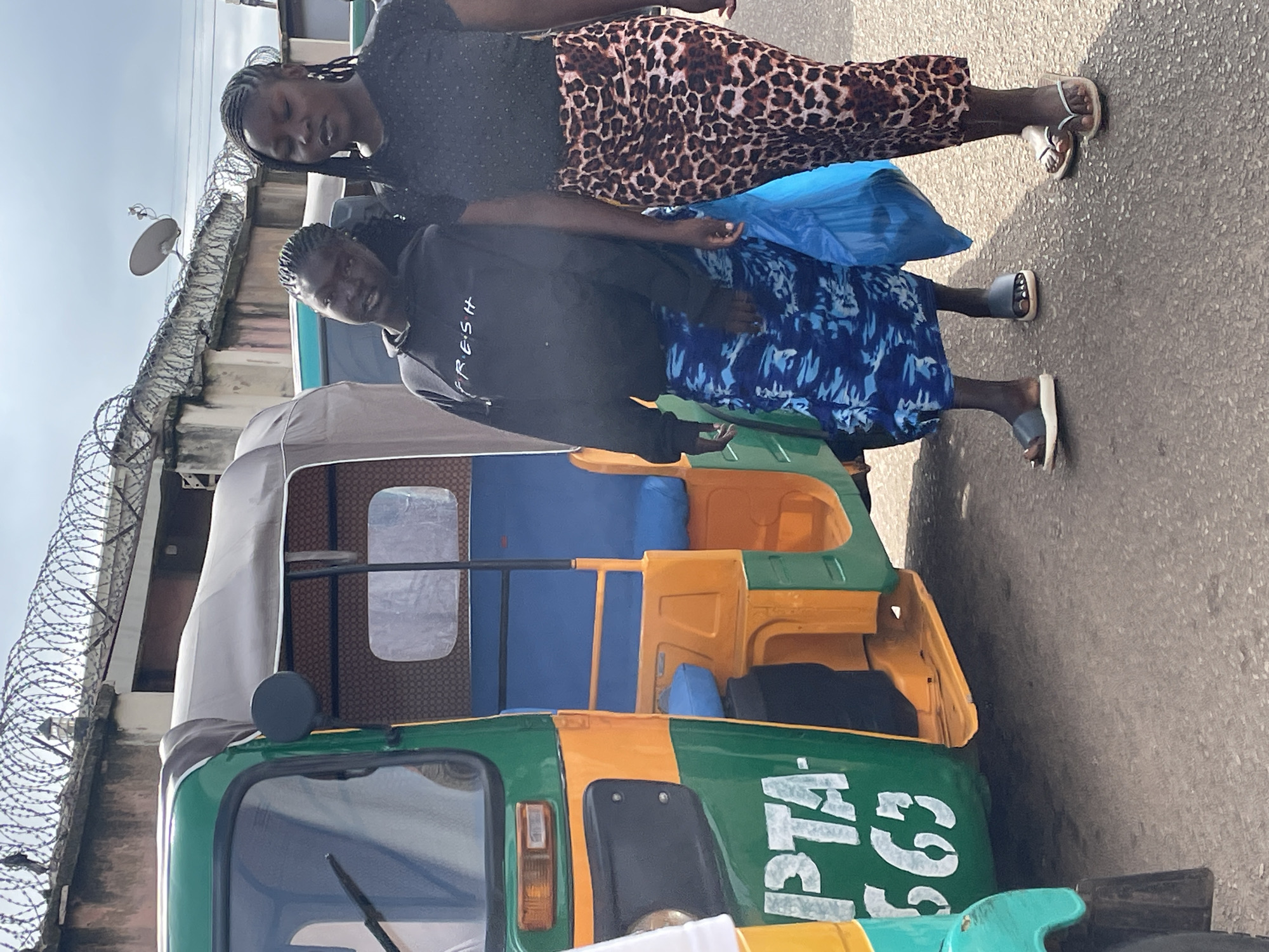 Two young Black women, one wearing a black hooded sweatshirt with "FRESH" in white lettering and a blue and white patterned skirt, the other wearing a black sparkly top and a leopard-print skirt, walk next to a green and orange auto-rickshaw, commonly known as an "okada" or "keke napep," on a paved road. The auto-rickshaw has "PTA 563" written in white lettering on its green side. Behind them, a concrete wall topped with barbed wire runs along the street, and a satellite dish is mounted on top of the wall. The sky is overcast. The scene appears to be in a bustling urban environment, consistent with locations in Jos, Nigeria, where auto-rickshaws are a common mode of transportation.