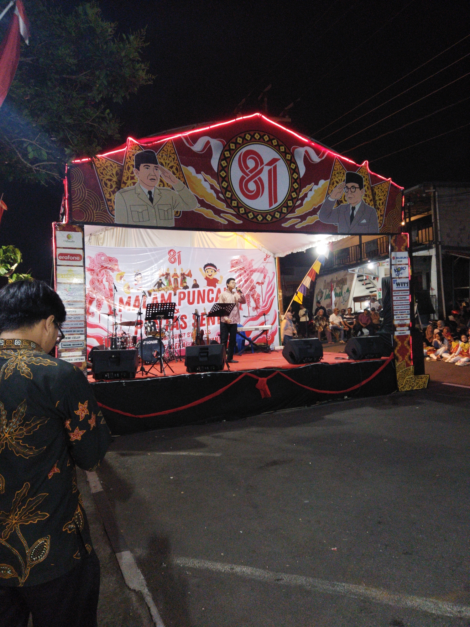 The image depicts an outdoor night-time event, likely a stage performance, in Semarang, Indonesia. A stage is set up with a large banner featuring portraits of two Indonesian leaders and the number "81" prominently displayed. The stage is decorated with red neon lights and traditional Indonesian motifs. A band with musical instruments, including drums and keyboards, is set up on the stage, and two individuals are seen performing, one singing into a microphone. A crowd of people is gathered in front of and around the stage, some seated on the ground, watching the performance. The foreground shows the back of a person wearing a batik shirt, looking towards the stage. The event appears to be taking place on a street, indicated by the asphalt road and white line markings. The overall atmosphere suggests a festive cultural gathering.