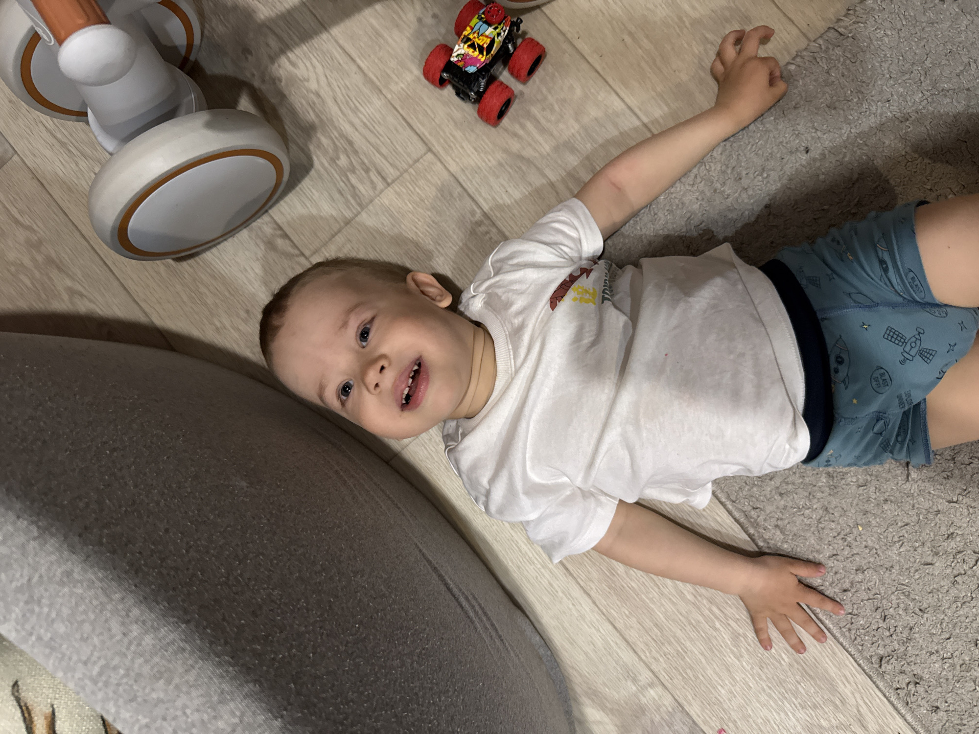 A young child is lying on their back on the floor. The floor is made of light-colored wood planks and a light gray carpet. The child is wearing a white t-shirt and blue shorts with a dark blue waistband. They are lying on their back with their arms outstretched. Next to the child is a red and black toy car. A light-colored toy walker with orange accents is visible in the background. The location is likely an indoor space, possibly a residence. Rusanovo, Russia, is not explicitly identifiable from the image.