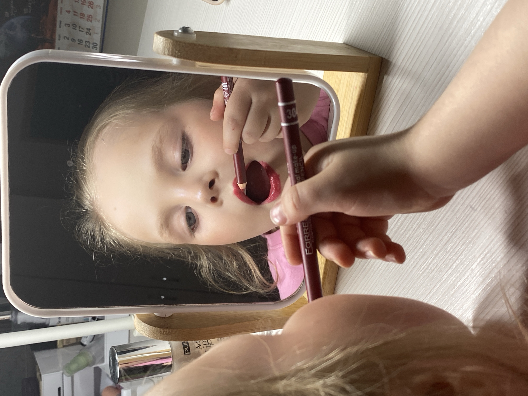 A young blonde child is captured in a close-up, reflected in a rectangular tabletop mirror set on a wooden stand, in what appears to be an indoor setting in Votkinsk, Russia. The child is actively applying dark red lip makeup using a pencil, with their mouth slightly open and tongue visible, demonstrating a focused and playful demeanor. The image is taken from just above the child's shoulder, looking towards the mirror, with the child's fair-skinned arm and hand holding the makeup pencil prominently in the foreground.
Visible text on the makeup pencil includes "FORRE" (likely a brand name), "al make-up PENCIL", and the shade number "30". Other items on the surface behind the mirror include a bottle with "PUR" and "Act" partially visible, suggesting cosmetic products. A calendar is also partially visible in the background reflection, showing dates such as "4", "9 10 11", "16 17 18 19", and "23 24 25 26", with a prominent "30" followed by "1 2 3", indicating the transition between months. The lighting is soft and even, suggesting an indoor, possibly daytime or well-lit evening, environment.