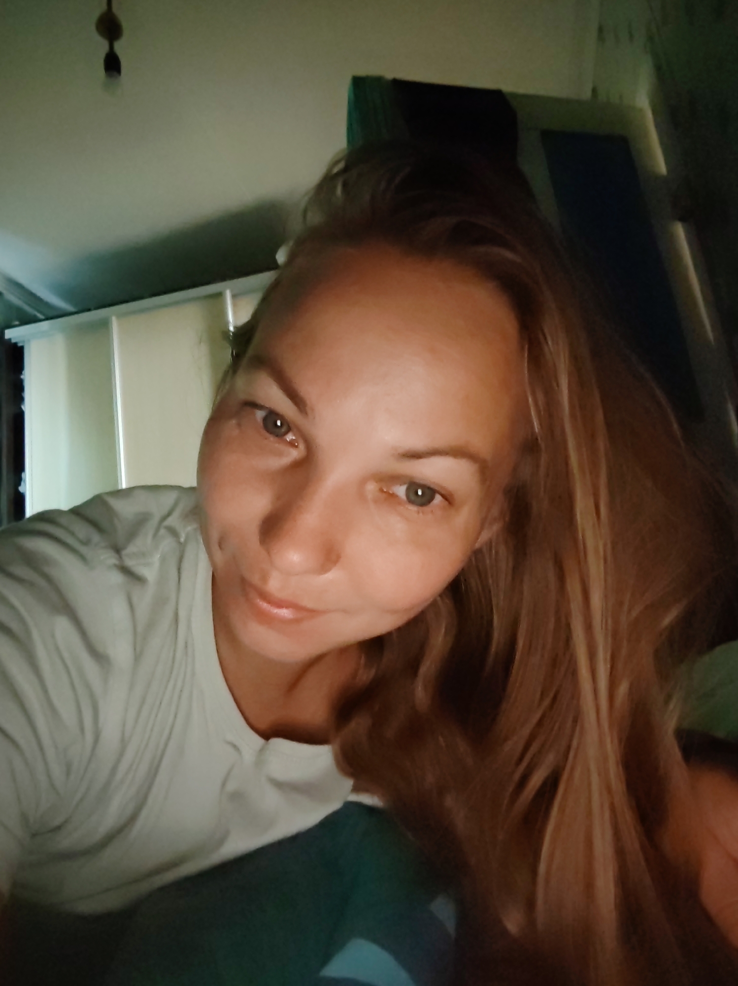 A close-up, head-and-shoulders selfie shows a woman with long, blonde hair. She is wearing a light-colored t-shirt and is looking at the camera. Her left eye is open and she has a slight smile on her lips. Dimples are visible on her cheeks. The background is dimly lit and appears to be an indoor setting with some shelving units visible. The lighting suggests it may be evening or the room is not well-lit. There is no indication of the specific location within the city of Чухлэм, Russia, but the environment appears to be a domestic or personal space.