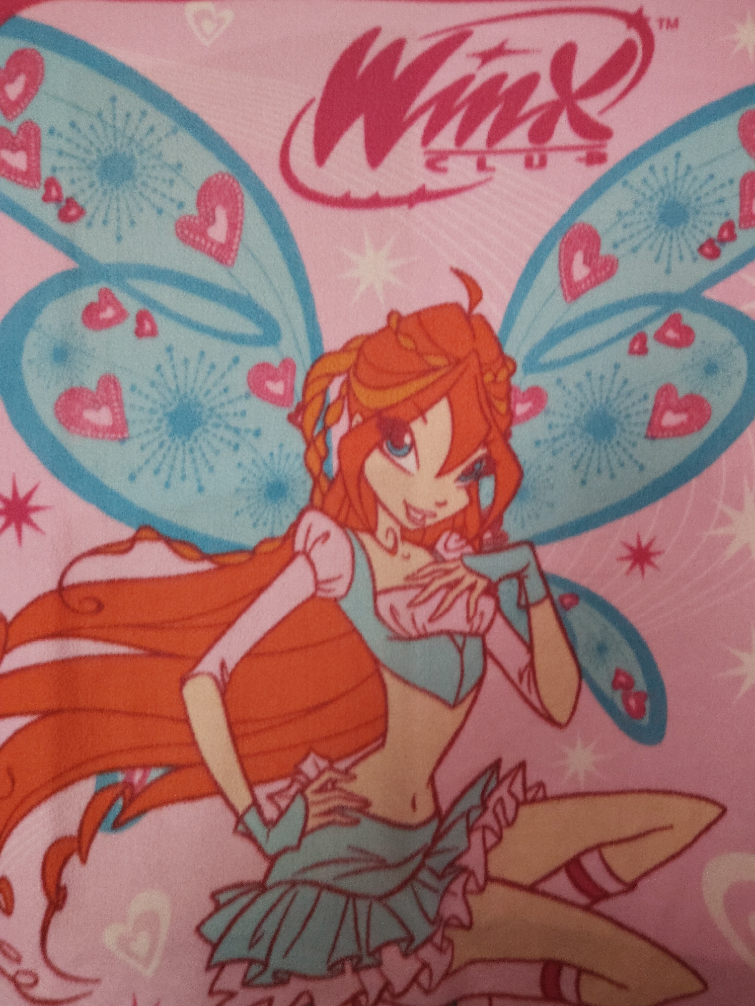 The image shows a cartoon rendering of Bloom, a character from the animated television series "Winx Club". She is depicted with long, red-orange hair, blue eyes, and fair skin. She is wearing a blue and pink outfit, and has large, blue wings decorated with pink hearts and white starburst patterns. The "Winx Club" logo is visible above her, in a stylized pink font. The background is pink and contains scattered small hearts and starbursts.