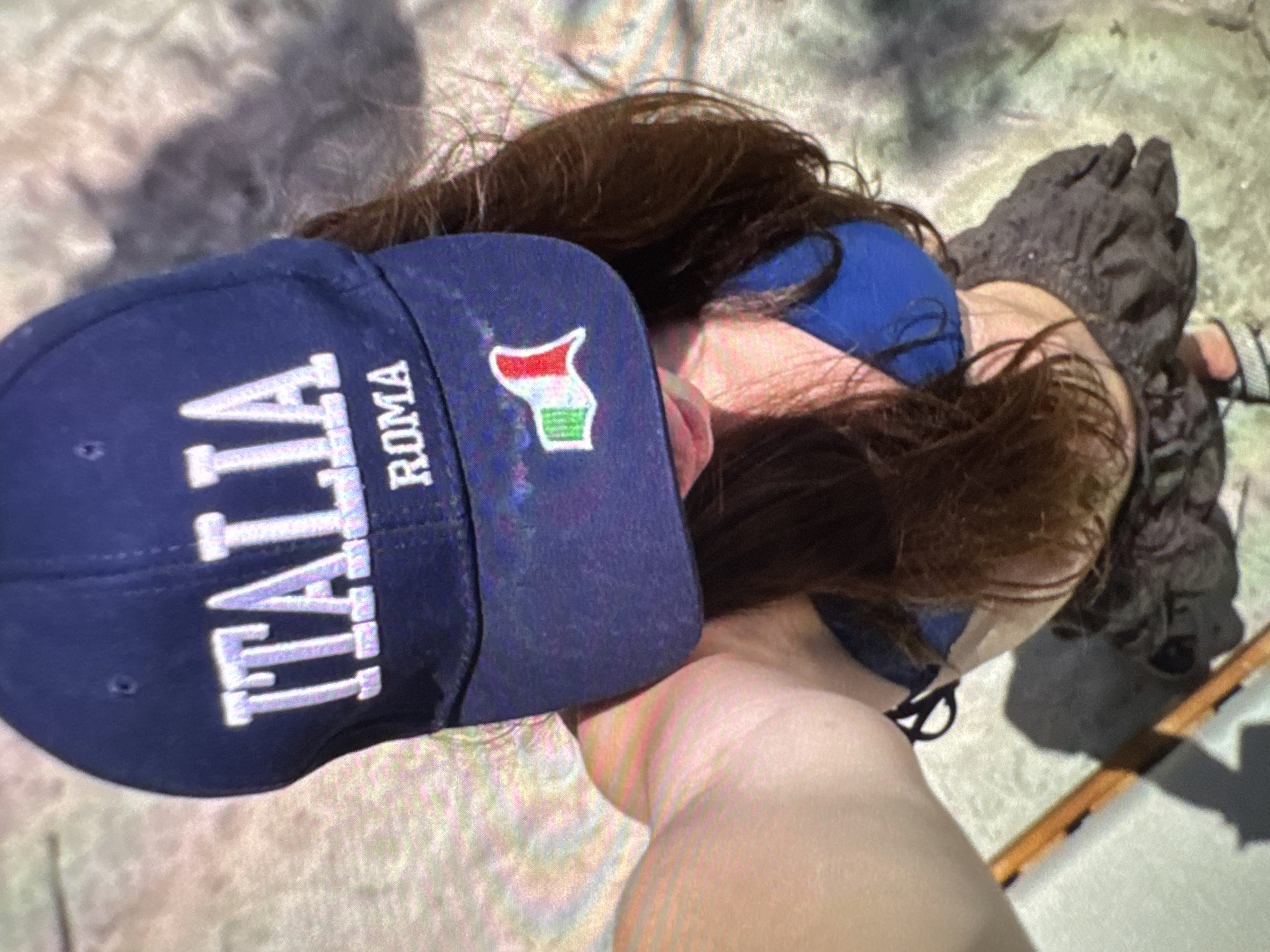 A person wearing a blue bikini and a blue baseball cap with "ITALIA ROMA" written in white is lying on a concrete surface in an unknown location. The cap also features a small emblem. The person has brown hair and is partially obscured by the cap. Their right arm is visible, extending towards the bottom of the frame. Next to the person is what appears to be a dark-colored bag or cloth. A portion of a wooden object is also visible.